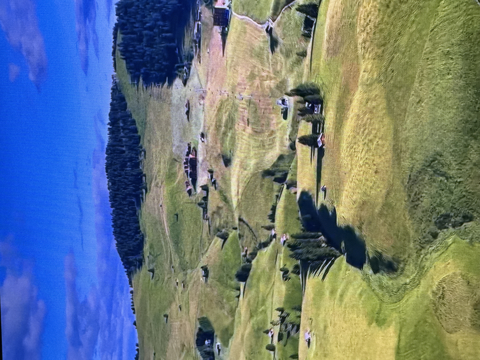 The image displays an aerial view of a mountainous landscape with rolling green hills and scattered trees. Several small buildings, appearing to be houses or cabins, are visible across the terrain. A ski lift or cable car system is present, ascending a grassy slope on a distant hilltop which is covered by a dense forest. The sky is a clear blue with a few wispy clouds. The scene is characterized by its natural beauty and rural setting. There is no indication that this location is in the city of Dubai, United Arab Emirates, as the landscape is not consistent with Dubai's geography.