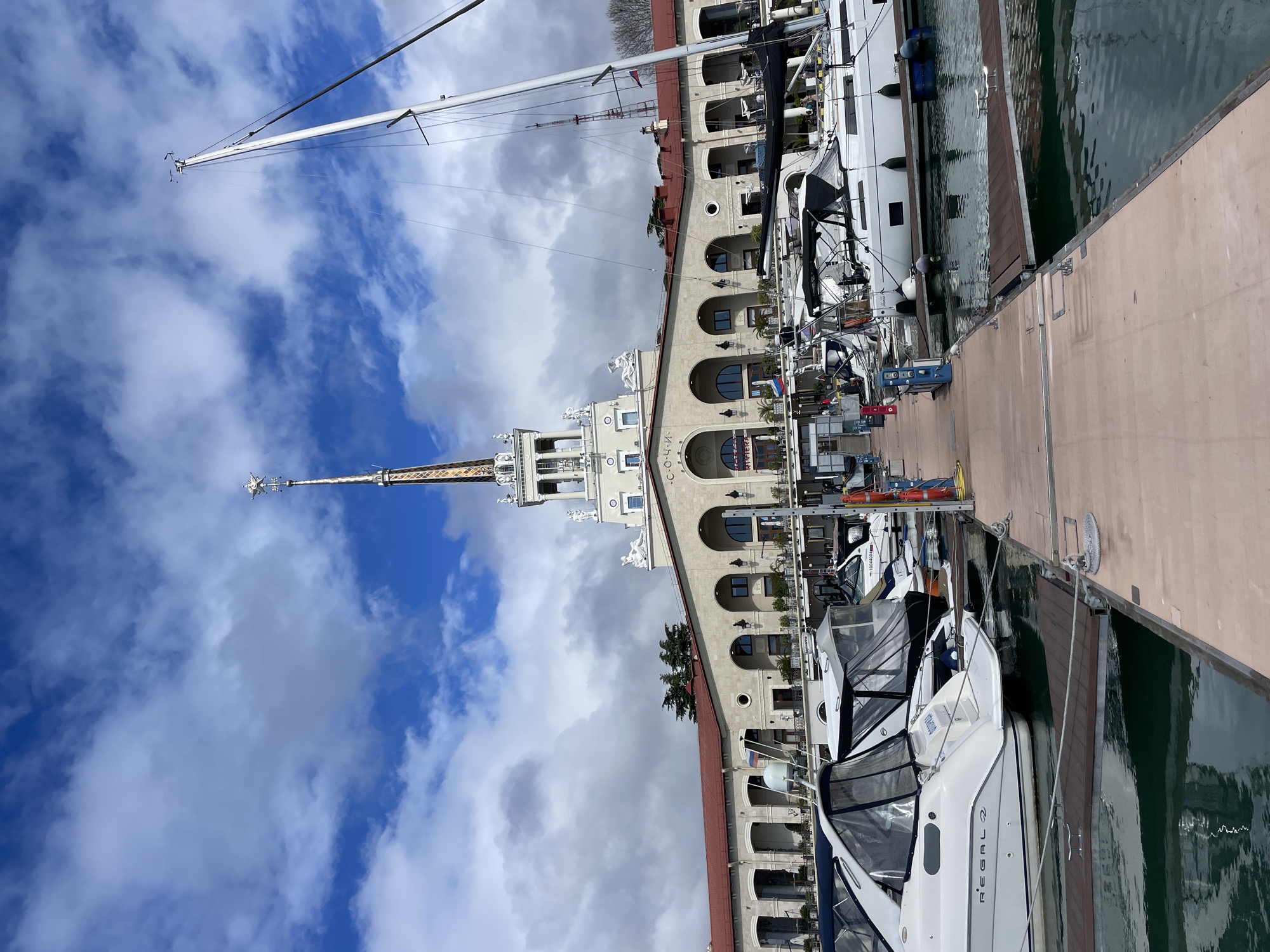 The image depicts a harbor scene in Sochi, Russia, prominently featuring the iconic Sochi Marine Terminal building. The building is characterized by its ornate architecture, including arches, statues, and a central spire that reaches into the sky. The harbor is filled with various types of boats, from small recreational crafts to larger yachts. The sky is filled with cumulus clouds and blue sky. The picture is taken on a sunny day.