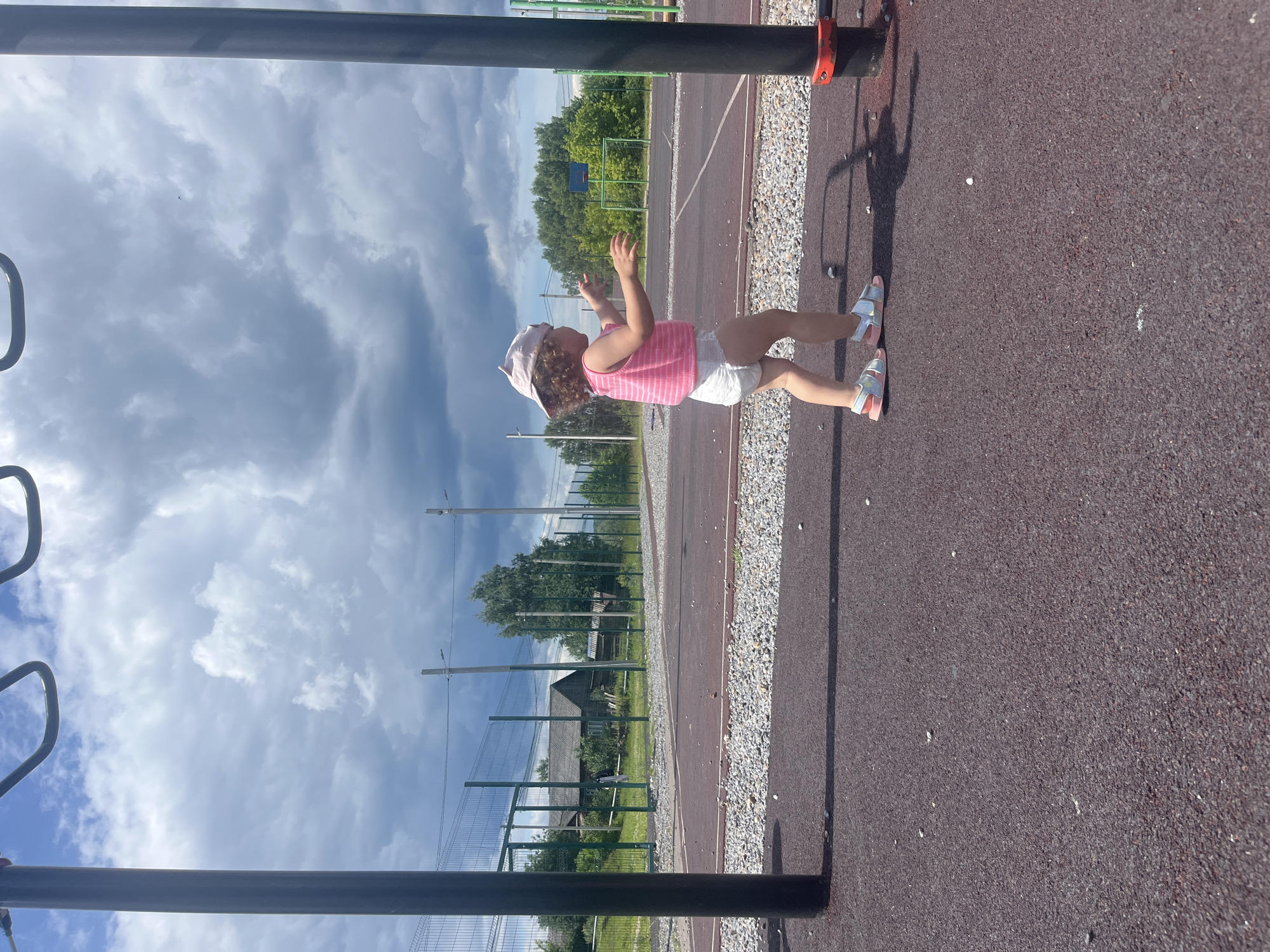A young girl wearing a pink striped tank top, white shorts, and a pink cap stands on a dark red, rubberized playground surface. She has on light pink and silver sandals. Her arms are raised, and she is looking up at the sky with an expression of wonder or surprise. The playground appears to be in a rural setting, with a chain-link fence and trees visible in the background. Beyond the trees, there are houses and a soccer goal. The sky is filled with dramatic, swirling clouds, suggesting either a storm is brewing or has just passed. The sun is out, casting a shadow of the girl and a metal bar in front of her onto the playground surface. The time of day appears to be late morning or early afternoon. There is no visible text in the image.