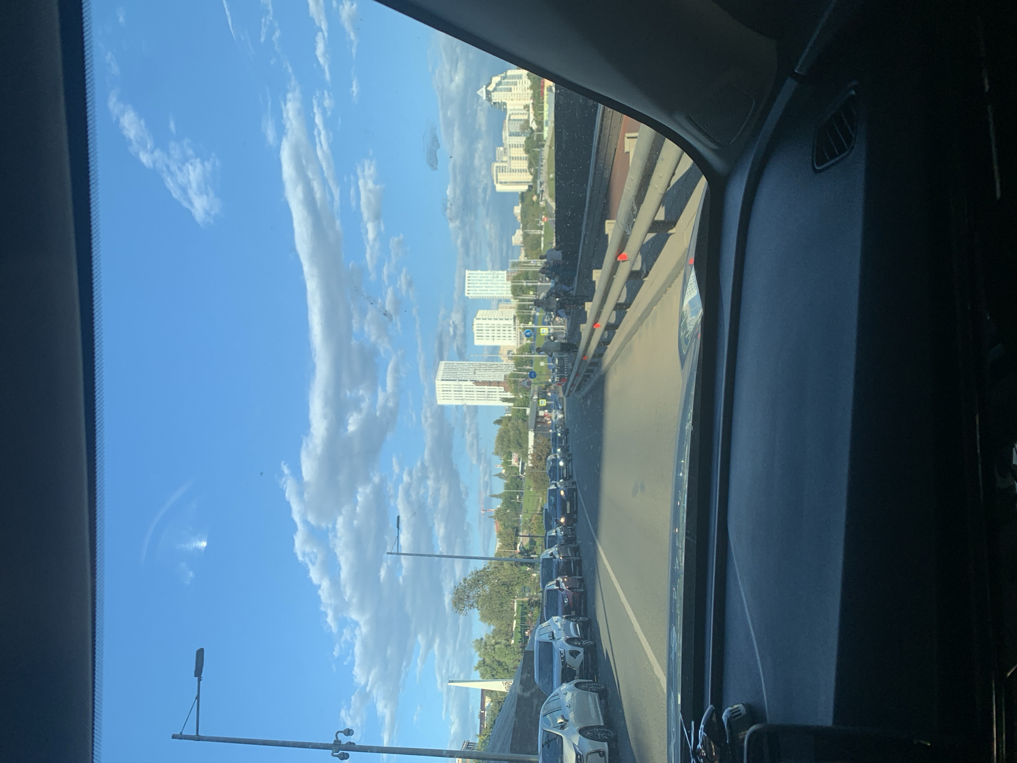 A car is stopped in heavy traffic on a multi-lane road, likely a bridge, in Ufa, Russia. The driver's perspective is visible, showing the dashboard and windshield. Outside the car, a line of vehicles is visible, stretching into the distance. Tall buildings, some modern high-rises, are present in the background, indicating an urban environment. The sky is blue with scattered white clouds, suggesting daytime. A street light is positioned on the left side of the road. There are some pedestrians visible further down the road.