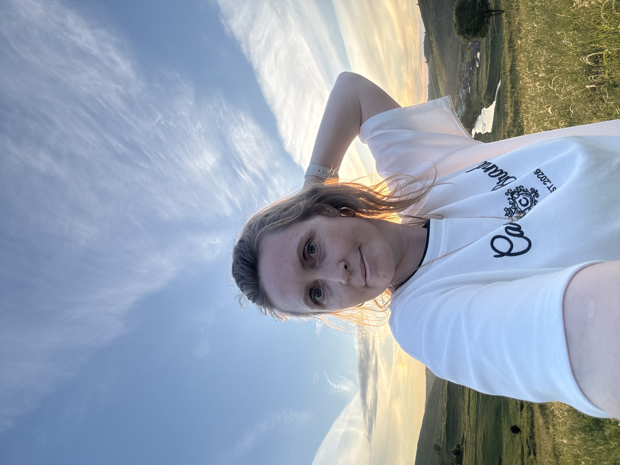 A woman with blonde hair pulled back is taking a selfie outdoors with her right arm raised behind her head, her left arm extended towards the camera. She is wearing a white t-shirt with black trim around the neckline and a gold chain with a pendant. The t-shirt has black text and a circular emblem. She has blue eyes and is looking at the camera with a slight smile. The background is a landscape with rolling green hills, tall grass in the foreground, and a river winding through a valley with scattered houses and trees. The sky is a clear blue with wispy white clouds, and the sun is setting, casting a warm glow over the scene. The photograph appears to have been taken in or near the city of Baiki, Russia, given the style of houses and the natural landscape.
