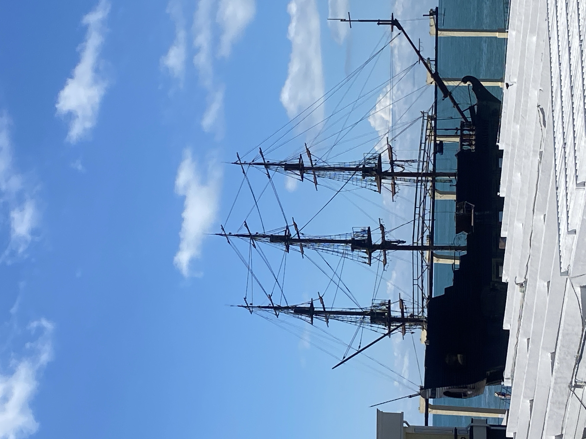 This is a vertical, eye-level, medium shot of a black, three-masted ship silhouetted against a bright blue sky with scattered white clouds. The ship is docked and appears to be stationary, possibly a museum or attraction. The masts are tall and feature rigging, but no sails are visible. The ship has a dark hull with a prominent bowsprit. In the foreground, there is a white, slatted structure, possibly part of a pier or walkway. The background shows a body of water that appears to be the sea. To the far left, a portion of a light-colored building is visible. A single person, wearing dark clothing and a hat, stands on a dock to the left of the ship. This image was taken in Sochi, Russia, likely near the city's waterfront or port area.