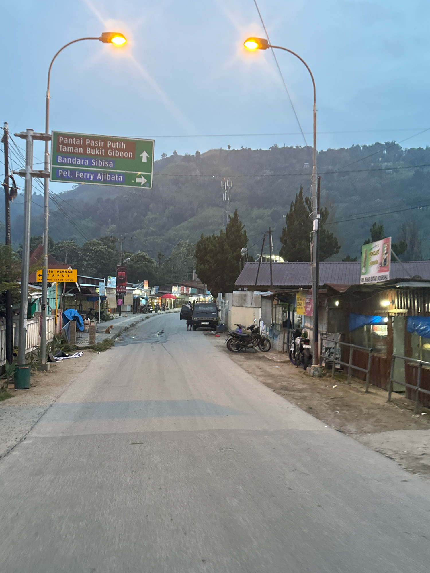 The image shows a street scene in Parsaoran Ajibata, Indonesia, likely during the early morning or late evening, as indicated by the illuminated streetlights and the soft, diffused light. In the foreground, the asphalt road is visible, with a light blue stripe painted across it, possibly a speed bump or a lane marker. The road is flanked by a dirt shoulder on the left and a row of small shops and buildings on the right.
On the left side of the road, a green directional sign points to "Pantai Pasir Putih, Taman Bukit Gibeon" (White Sand Beach, Gibeon Hill Park) with an upward arrow, and "Bandara Sibisa" (Sibisa Airport) and "Pel. Ferry Ajibata" (Ajibata Ferry Port) with rightward arrows. A smaller yellow sign with Indonesian text is also visible, with an arrow pointing left. Power lines and poles are present, along with a green trash can.
The shops on the right are simple structures with corrugated metal roofs. Various items are displayed outside, including what appear to be drinks and other goods. Several motorcycles are parked along the roadside. A dark-colored pickup truck is parked near one of the shops, with its door open. A small, light-colored dog is sitting on the ground on the left side of the road.
In the background, a hill covered in trees rises, with a few scattered buildings visible on its slopes. A cellular tower is also present on the hillside. The sky is overcast, contributing to the diffused lighting. The overall impression is of a functional, somewhat rustic, local street serving as a connection point for various destinations.