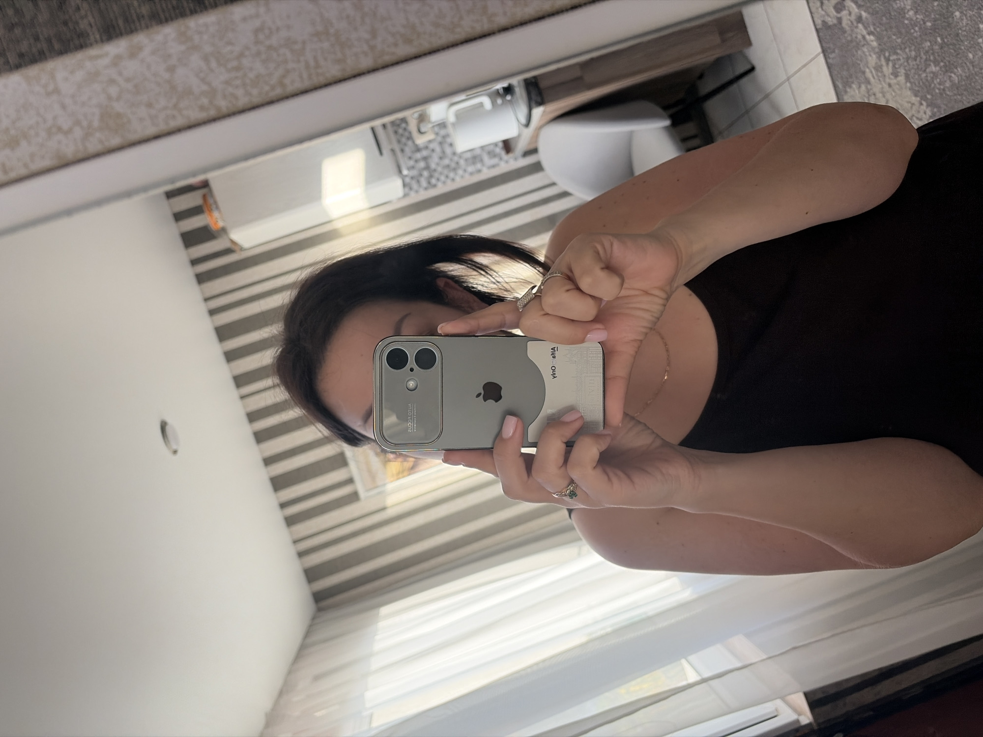 The image is a mirror selfie, rotated 90 degrees counter-clockwise. A person, appearing to be female, is shown holding a smartphone to capture their reflection. The person is wearing a dark top, with dark hair partially visible around the phone. Both hands are holding the phone; the left hand (right in the reflection) has a gold-colored ring with a green stone on the ring finger, and the right hand (left in the reflection) wears a silver-colored ring with small clear stones on its ring finger. Her nails have a light pink or nude polish. A delicate gold-colored chain is visible around her neck.
The smartphone is an iPhone with a distinctive triple-camera setup, featuring a silver/gray back and a visible Apple logo. It is encased in what appears to be a clear or translucent case. Mirrored text is visible on the phone case, including "HOA YЯTJU" on the right side and other characters (e.g., "2100d OUI") on the left side of the reflection, likely part of the case's branding or features.
The background, reflected in the mirror, shows an indoor setting, likely a room in Sochi, Russia. A prominent feature is a wall covered with horizontal light and dark gray stripes. A window with white sheer curtains or blinds allows natural light into the room, suggesting it is daytime. Further back in the reflection, a light-colored object resembling a chair or toilet and a counter or shelf with small white objects are discernible. A textured, light-colored rug or floor covering is also visible in the upper part of the reflection. A recessed light fixture is seen on a white ceiling.
The activity is the person taking a mirror selfie. The scene depicts a casual, personal moment in an interior space.