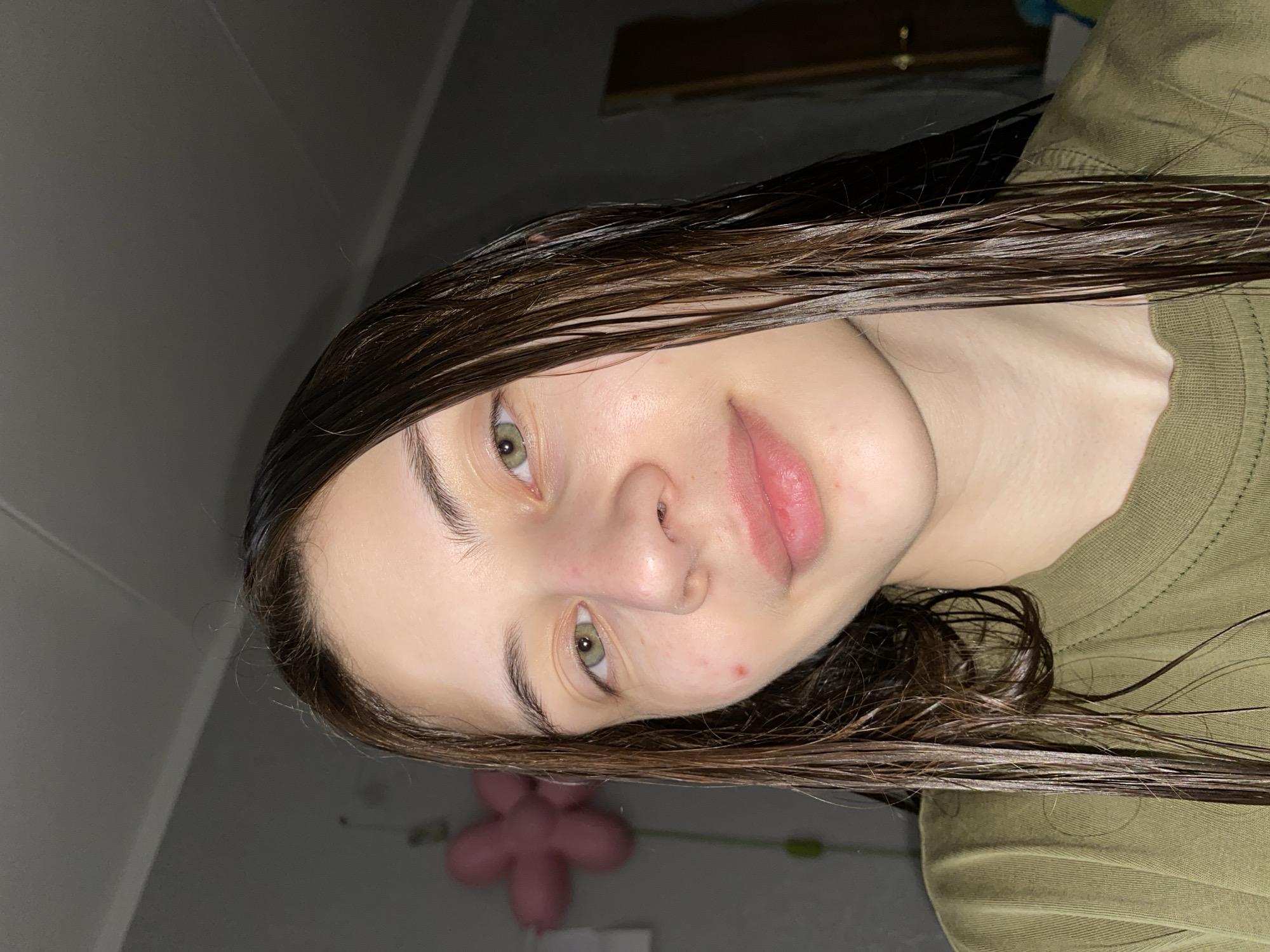 A close-up, slightly angled shot of a young woman with wet, dark hair. Her skin appears pale and clear, with a few small blemishes on her face. She has light-colored eyes and pink lips. She is wearing a muted green top. The background includes a light grey wall and a partially visible pink balloon decoration. There is no evidence to suggest this image was taken in Votkinsk, Russia.