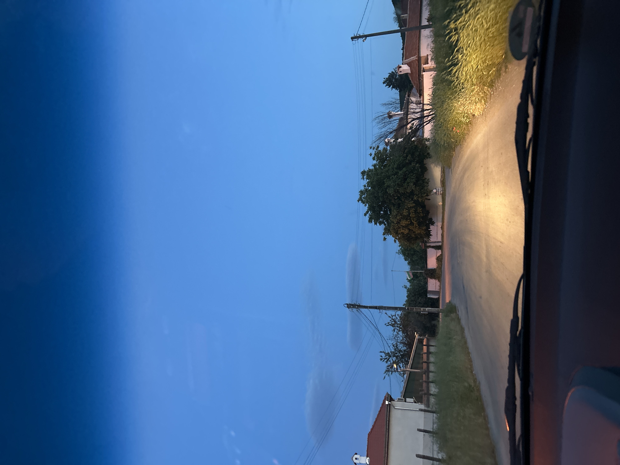 The image shows a view from inside a vehicle driving on a road in Ovchepoltsi, Bulgaria, at dusk. The sky is a gradient of blue. Buildings and trees line the side of the road. Telephone poles with wires run parallel to the road. The vehicle's windshield wipers are visible. The road is illuminated by the vehicle's headlights.