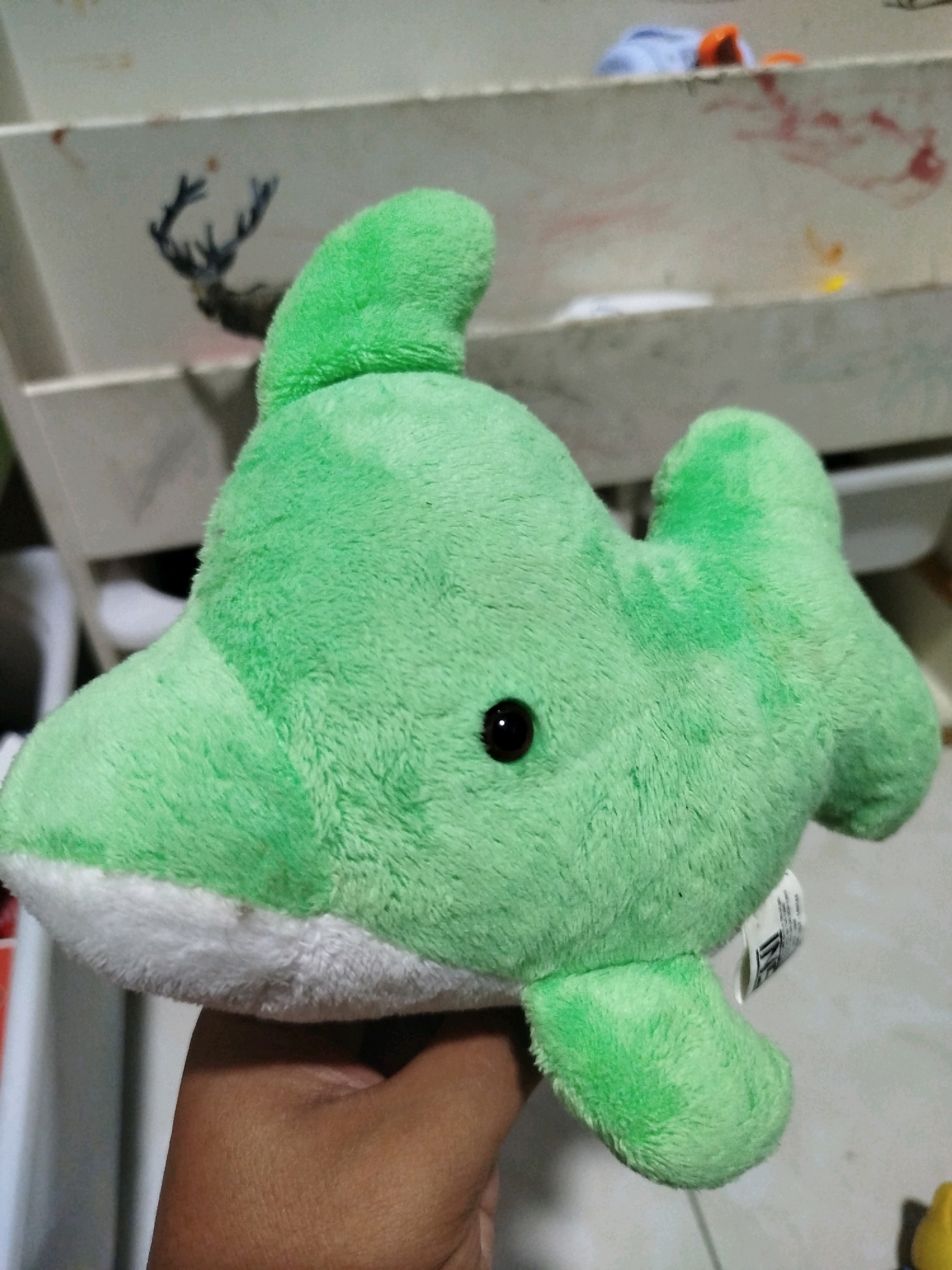 A plush green dolphin toy is being held by a person's hand. The toy has a white underside, a black eye, and two fins on its back. The background consists of a light-colored, multi-tiered surface with some smudges and what appears to be a drawing of a deer's head. The setting is likely indoors, possibly a room or workspace, given the presence of shelves and miscellaneous items in the background. The image was captured in Mojoagung, Indonesia.