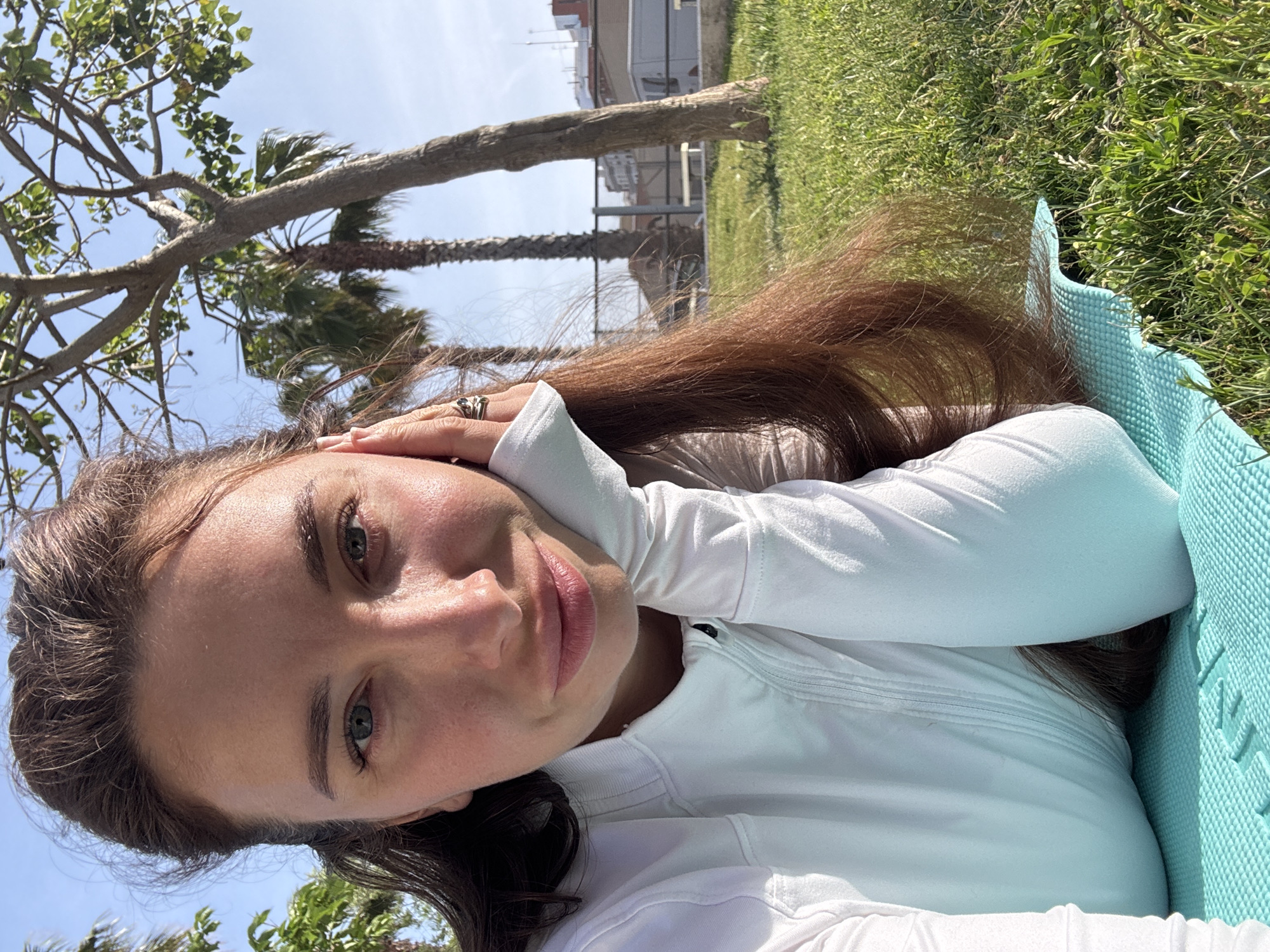 The image is a bright, sunlit, selfie-style portrait of a young woman lying outdoors on a light blue mat. She is positioned on her side, looking directly at the camera with a calm, neutral expression. She has long, dark brown hair, fair skin, and light-colored eyes. She is wearing a white long-sleeved athletic or casual top. Her left hand, adorned with a ring featuring dark stones, is resting near her head.
The setting is an outdoor green space, likely a park or garden, bathed in clear, bright daylight. Lush green grass surrounds the mat she is on. Above her, the branches and green leaves of a large deciduous tree are prominent against a clear blue sky. In the mid-ground, several palm trees are visible. Beyond the trees, a modern building with balconies can be seen, hinting at an urban or semi-urban environment.
The time of day appears to be midday or early afternoon due to the strong, direct sunlight and lack of long shadows. The weather is clear and sunny. The overall atmosphere is serene and relaxed. On the light blue mat, faint embossed text is visible along its edge, with discernible letters including "INE," possibly part of a brand name or product type like "YOGA LINE." The palm trees and architectural style align with the given location context of València, Spain.