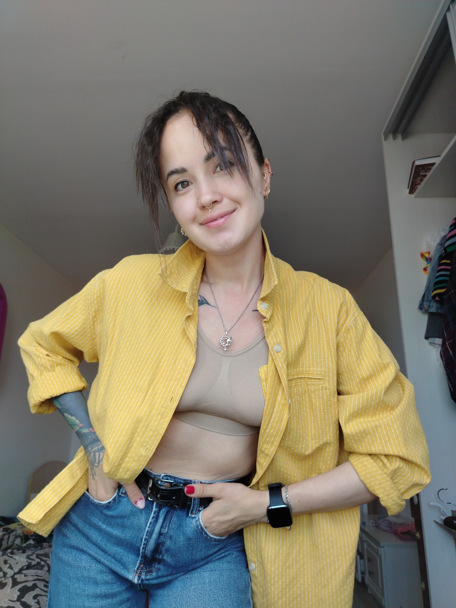 A woman with dark hair and a nose ring smiles at the camera, wearing a yellow pinstripe shirt, a beige bralette, and blue jeans. Her left arm, which is tattooed, is resting on her hip, and her right hand is in her jeans pocket. She is also wearing a silver necklace, earrings, and a black smartwatch. The scene appears to be indoors, with a closet visible in the background. The lighting suggests it might be daytime.