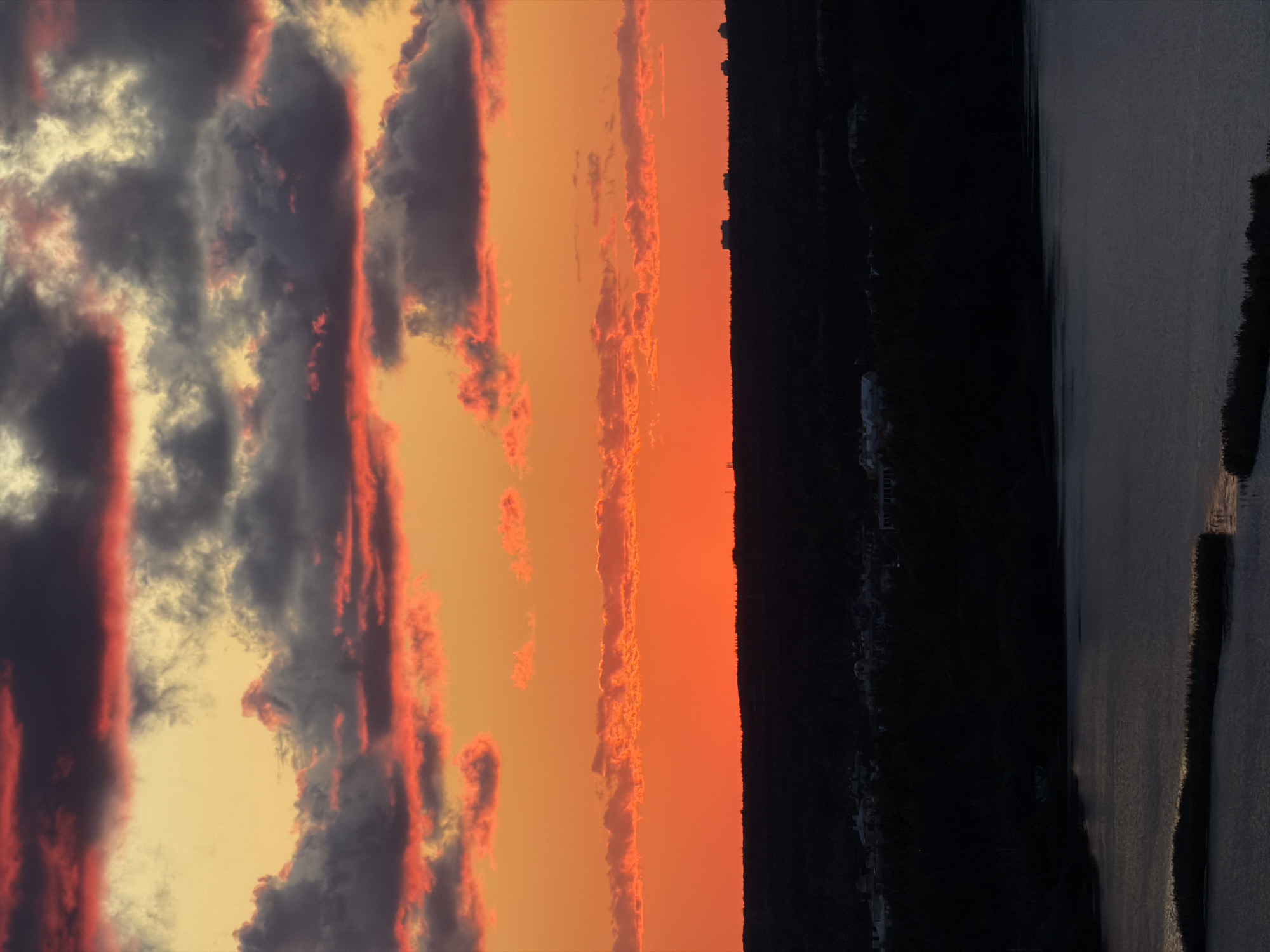 The image depicts a vibrant sunset over a body of water and a dark, silhouetted landscape. The sky is filled with dramatic clouds, painted in hues of orange, red, and yellow, with streaks of dark grey and deep red highlighting the edges of the clouds. The horizon is a dark, undulating line, suggesting a densely wooded area or hilly terrain. A body of water, appearing calm, occupies the lower portion of the frame, reflecting the subdued light from the sky. Small, indistinct structures are visible along the distant skyline. The overall impression is one of natural beauty and tranquility, a common sight in the outskirts of Saint Petersburg, Russia, particularly during the evening hours when the sun sets behind the city's green belts and adjacent bodies of water. The specific location within Saint Petersburg is not identifiable from the provided image.