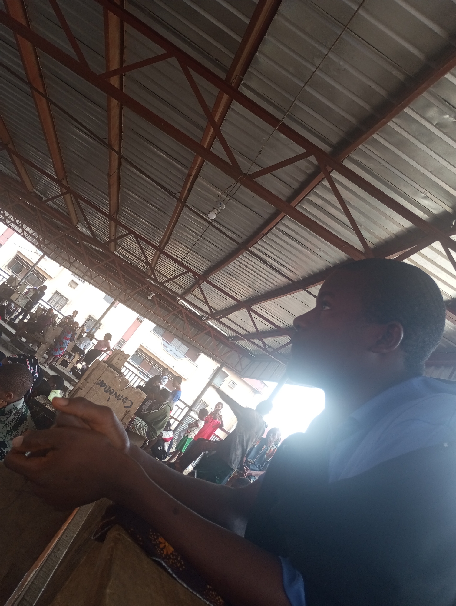 The image captures a close-up of a person's forearms and hands, clasped together, in the foreground. The person appears to be sitting or standing within an open-air market setting in Onitsha, Nigeria, likely under a corrugated metal roof supported by a metal frame, as evidenced by the ceiling structure and lighting fixtures visible. In the blurred background, a bustling market scene unfolds with several individuals, stalls, and building structures. A prominent cardboard box with the word "Convention" written on it is visible. The lighting is uneven, with bright sunlight streaming in from the left, creating lens flare and silhouetting some figures in the background. The overall impression is of a busy public space, possibly a market or gathering area.