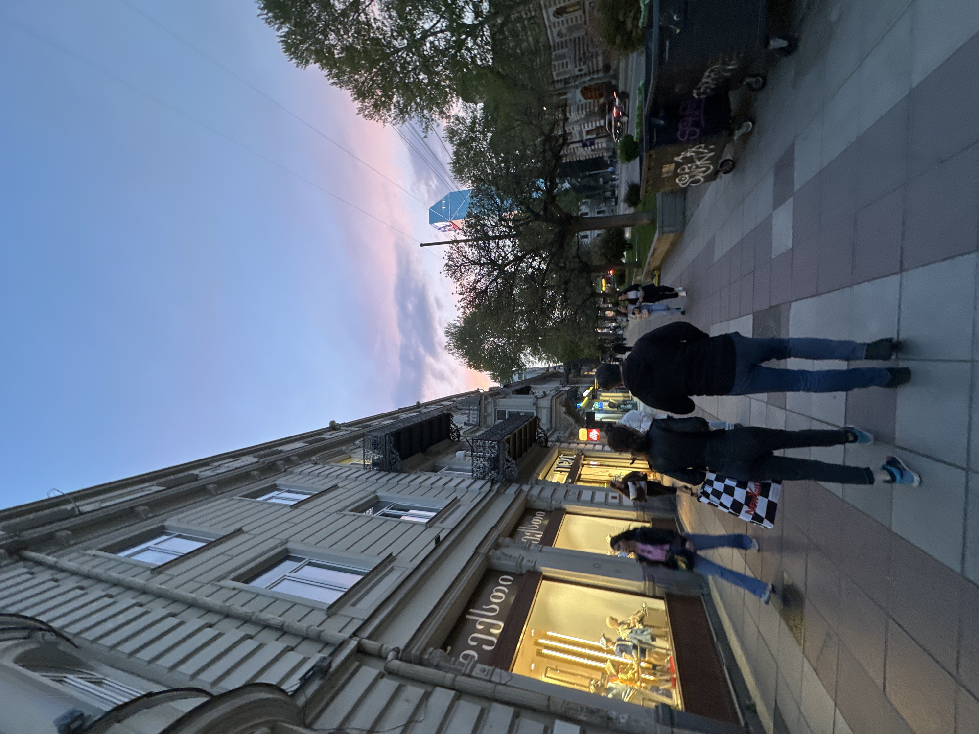 The image captures a vibrant city street scene at dusk, likely in Tbilisi, Georgia, given the visible Georgian script. The sky displays a gradient of soft blue transitioning to pink and purple clouds, indicating early evening. Overhead power lines crisscross the upper part of the frame.
On the left, a multi-story building with a light-colored facade, ornate balconies, and numerous windows lines the street. Its ground floor features brightly lit storefronts. Several pedestrians are walking along the wide sidewalk, which is paved with alternating light and dark gray squares. One person in the foreground wears a dark jacket and blue jeans, while another carries a distinctive black and white checkered shopping bag. Further down the street, more individuals are visible, some standing near storefronts, suggesting shopping or browsing.
In the mid-ground, mature trees with lush green foliage partially obscure the view of the street beyond. Peeking above the trees in the distance is a striking, modern-looking blue cuboid structure, possibly a building or artistic installation, with a grid-like pattern.
Visible text includes "ჯეოსელი" (Jeoseli, a Georgian word) on one storefront, "NEXT" on another, and "illy" further along. Graffiti, including the word "SUKA," is visible on a utility box or wall on the right side of the street. A vehicle can be faintly seen driving in the background on the road adjacent to the sidewalk. The overall atmosphere is that of a bustling urban environment during the golden hour.