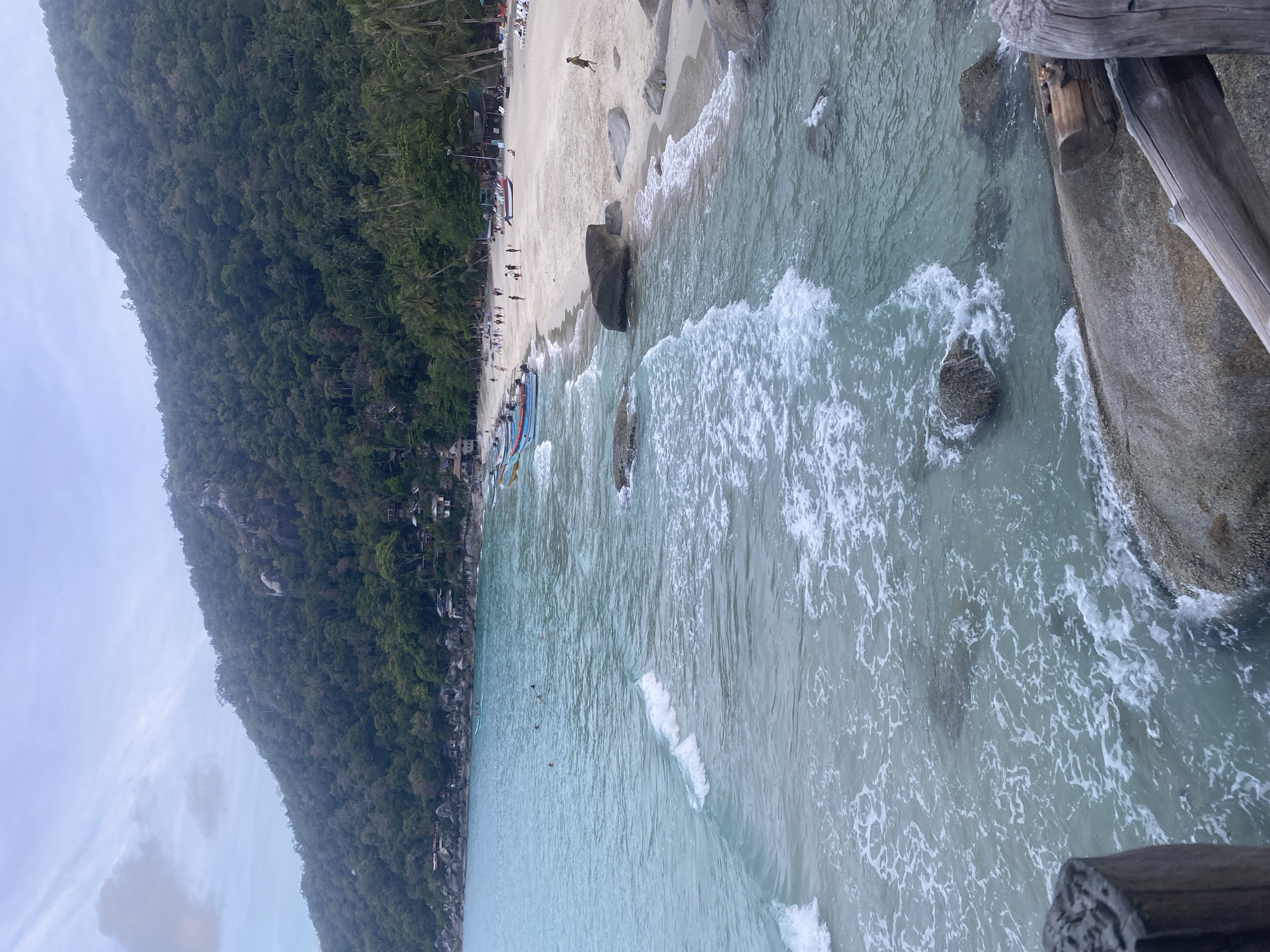 The image depicts a coastal scene in Ban Haad Rin, Thailand. A densely vegetated hillside dominates the left side of the frame, bordering a sandy beach where several people are present. Some people are walking along the beach, while others appear to be swimming in the turquoise water. Numerous boats are beached along the shore. In the foreground, the edge of a wooden structure, possibly a pier or walkway, is visible, with rocks and turbulent water. Waves are crashing against the shore and rocks. The sky is overcast, suggesting a cloudy day.