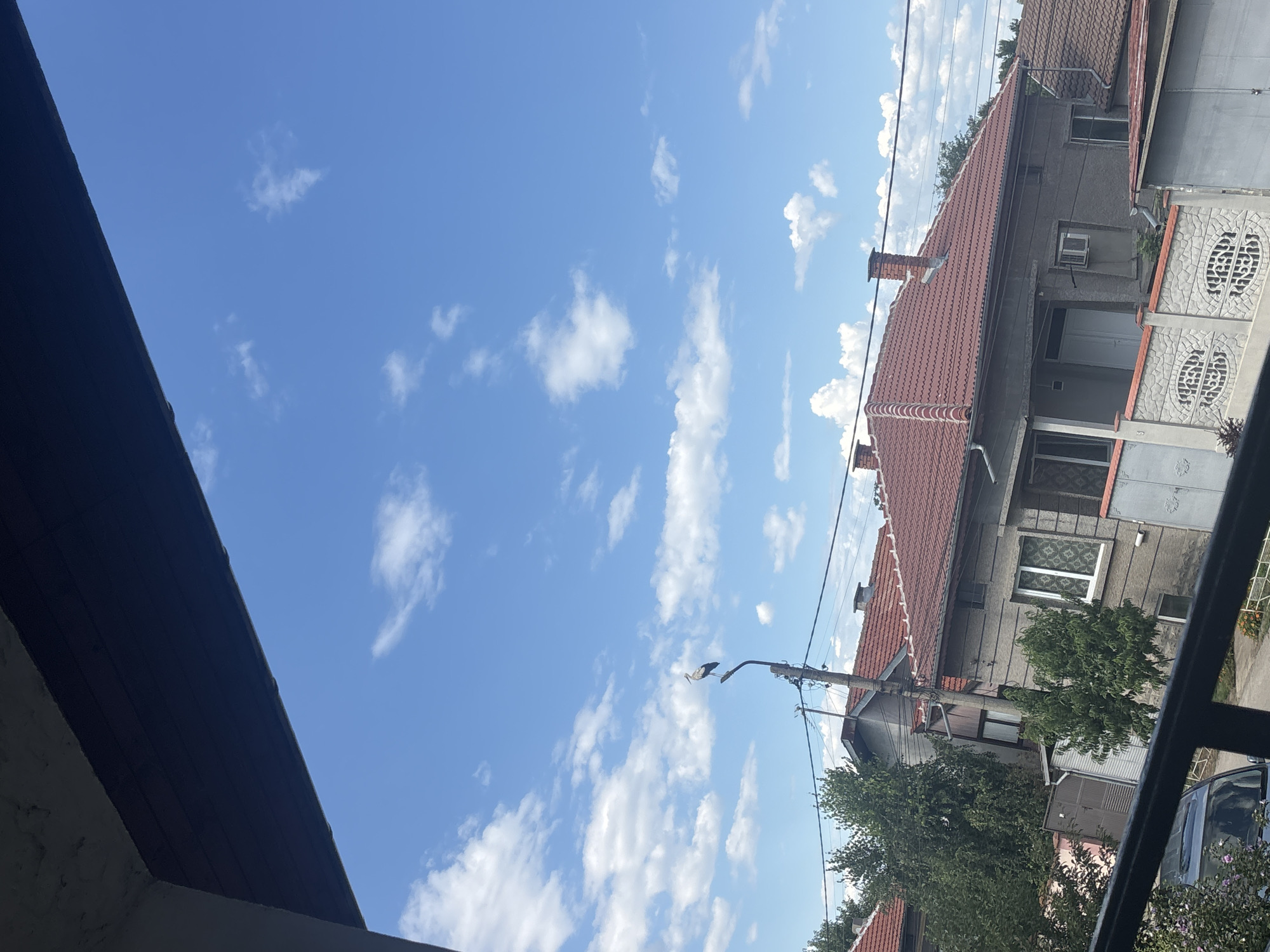 The image depicts a residential area under a partly cloudy blue sky, likely in the city of Marten, Bulgaria. A stork is perched on top of a utility pole in the mid-ground. In the foreground, a dark balcony railing is visible, suggesting the photo was taken from an elevated position. Behind the pole, houses with red tile roofs and gray facades are situated. Some of the houses have visible windows, doors, and chimneys. Trees are interspersed between the buildings, and a fence with decorative patterns runs along the front of one property. The overall scene suggests a typical suburban or town environment.