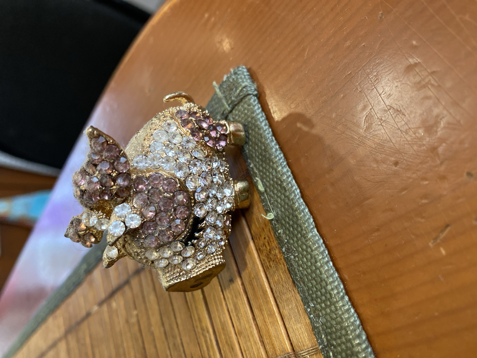 A jeweled pig figurine is the primary subject of this image, resting on a surface that appears to be a wooden instrument. The pig is encrusted with both clear and pinkish-purple rhinestones, giving it a sparkly and decorative appearance. It has small gold-colored ears and what seem to be wings, also adorned with the same gemstones. The figurine is positioned diagonally across the frame, with its head pointing towards the left and its body extending towards the right.
The surface beneath the pig is made of wooden slats, possibly part of a musical instrument like a lute or a guitar's soundboard, given the curvature. A strip of textured, green fabric runs along the right edge of the wooden surface, bordering the pig and partially overlapping it. The background is softly blurred, with a dark area on the left and a warm, brown wooden tone dominating the upper right. The lighting suggests it's daytime, with soft light casting subtle shadows. The overall impression is a close-up shot of a decorative item placed on a unique surface. There is no visible text in the image.