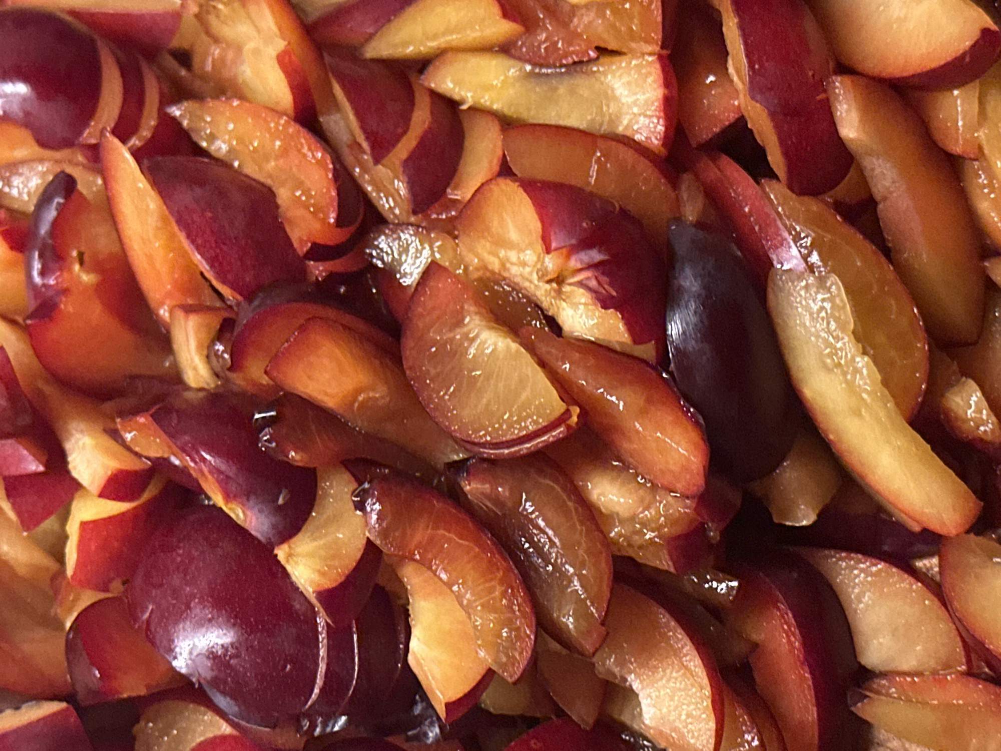 The image is a close-up, overhead shot of chopped plums. The fruit is cut into wedges and pieces, revealing the deep red skin and bright orange flesh. The plums appear juicy and are arranged in a dense, overlapping pattern filling the frame. The lighting is warm and highlights the glossy texture of the fruit. There is no discernible environment beyond the fruit itself, and no people or actions are depicted. The image does not contain any information that would allow for its location to be identified in the city of Nevinnomyssk, Russia, or anywhere else.