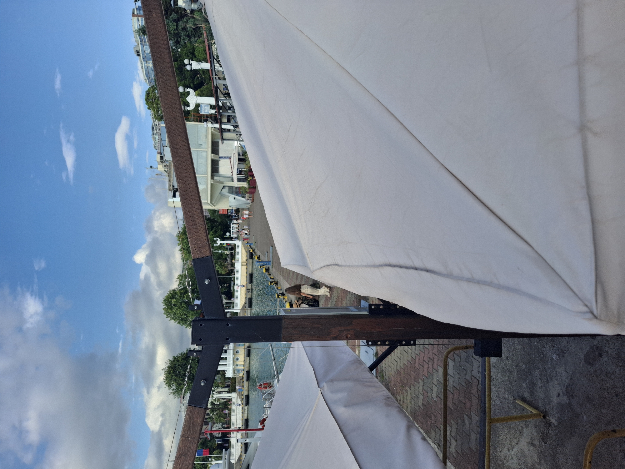 The image captures a scene from Sochi, Russia, likely during the daytime given the bright sky and clear visibility. The perspective is from underneath a large white canopy or umbrella, looking outwards towards a waterfront area. The canopy dominates the foreground, its fabric creating diagonal lines across the frame.
In the background, a bustling waterfront is visible. Buildings line the shore, with multiple stories and light-colored facades, suggesting a commercial or residential area. Trees are interspersed between the buildings and along the waterfront, adding a touch of greenery. The water itself appears calm, with a few boats or vessels docked along the piers. There are people visible in the distance, walking along the waterfront promenade. Some are dressed in casual clothing.
The structure supporting the canopy is made of dark wood and black metal brackets, with visible bolts and chains connecting parts of it. The ground in the immediate foreground appears to be concrete or paving stones. The sky is a vibrant blue, with scattered white clouds.
Notable details include the clear blue sky indicating good weather, and the architecture of the buildings which suggests a European style, consistent with a location like Sochi. The overall impression is of a pleasant day at a seaside resort or a lively waterfront district. There is no visible text in the image.