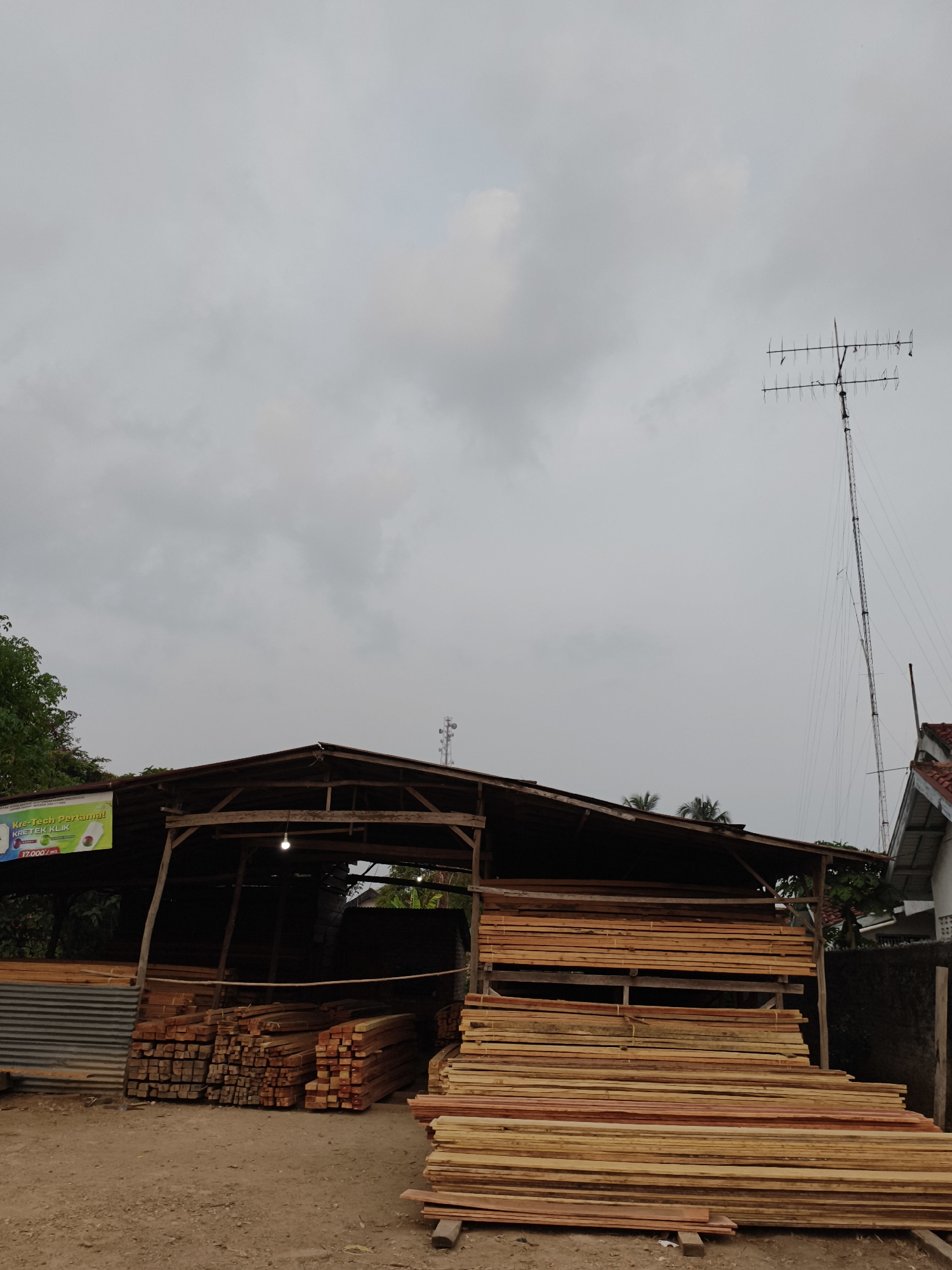 A medium shot captures an outdoor lumberyard in Kemala Raja, Indonesia, under a heavily overcast sky. The scene is dominated by stacks of lumber, both rough-cut and processed. To the left, a simple wooden structure with a corrugated metal roof shelters additional lumber and a metallic corrugated wall. A banner, partially visible on the left, advertises "Kre-Tech Pertama! KRETEK KLIK" with prices. In the center and right, large piles of wooden planks are neatly stacked. Behind the lumberyard, a tall, slender radio tower with multiple antennas extends towards the sky, and a portion of a residential building with a tiled roof is visible on the far right. The ground is dirt and gravel. No people are visible in the frame.
