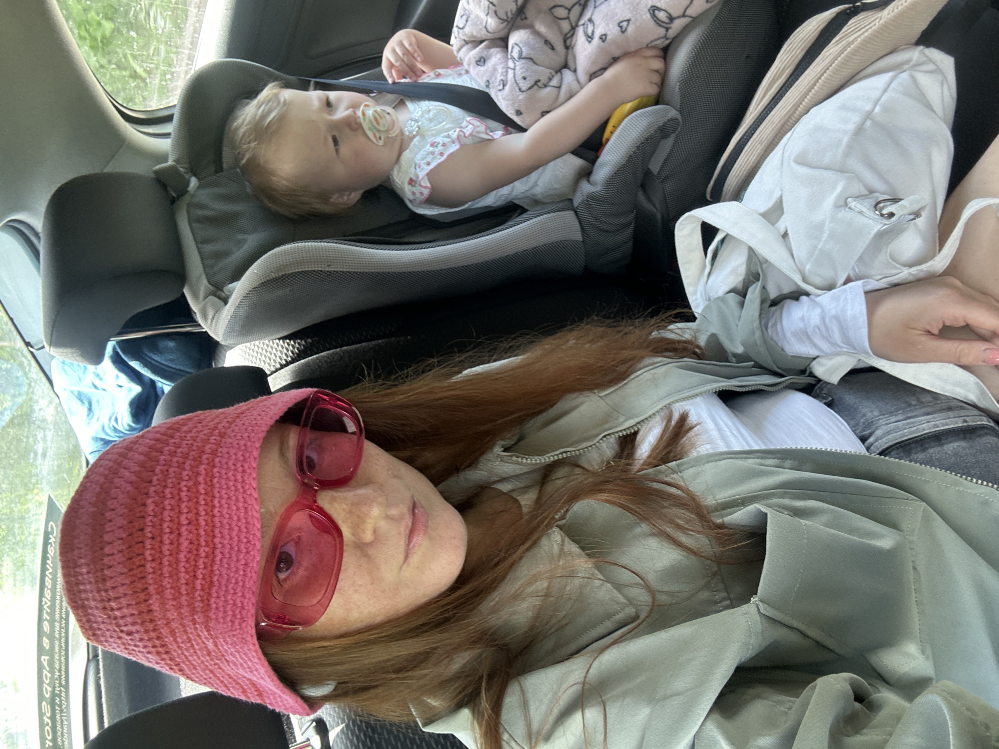 The image captures an angled, overhead view inside a car, featuring a woman and a baby. The woman, with long reddish-brown hair, is in the foreground, wearing a vibrant pink knitted headscarf and matching pink rectangular sunglasses. She has a light green/grey jacket over a white long-sleeved top and appears to be wearing dark trousers or jeans. Her expression is neutral as she looks towards the camera.
Behind her, positioned in a rear-facing grey and black car seat, is a baby or toddler with light blonde hair. The baby is dressed in a white top with a small floral pattern and has a pacifier in its mouth. A light-colored blanket with a faint pattern is draped over the baby, partially covering its body.
To the right of the woman, on the car seat, rests a white fabric bag with a metal ring, and a beige zippered bag or pouch. Bright daylight illuminates the car's interior, filtering through the windows, suggesting it is daytime. Outside the car windows, green foliage is visible, indicating a natural outdoor setting. A notable detail is the reversed, blurred Cyrillic text reflected on the window next to the woman's head. Parts of the text "CKAHHBAHHE" (SCANNING) and "B 9T OAA" are somewhat discernible, suggesting a sign or advertisement outside. The scene depicts a car journey in Nizhny Novgorod, Russia.