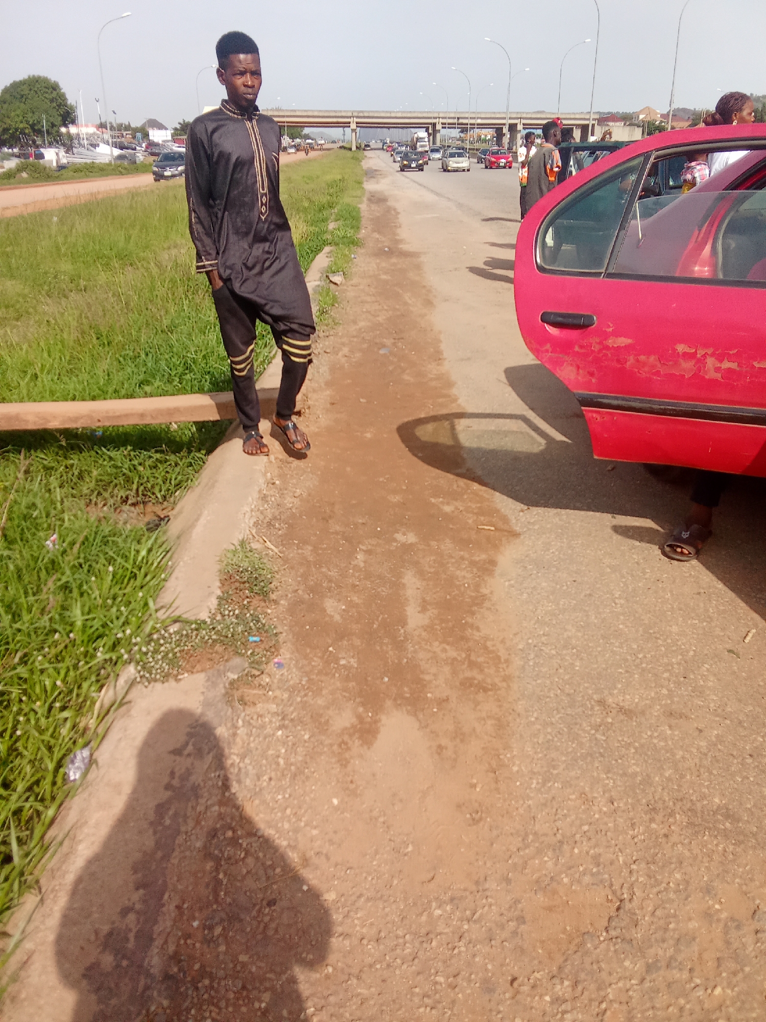 A full-body shot shows a young Black man standing on a dusty road in Kagini, Nigeria, next to a red car with its door open. The man is wearing a black traditional outfit with gold trim and brown sandals. His left hand is in his pocket, and he is looking to his left. His shadow is cast on the ground in front of him. The red car's paint is chipped and faded. In the background, there is a highway with cars and trucks, an overpass, and lush green grass on the left side of the road. The sky is clear and blue.