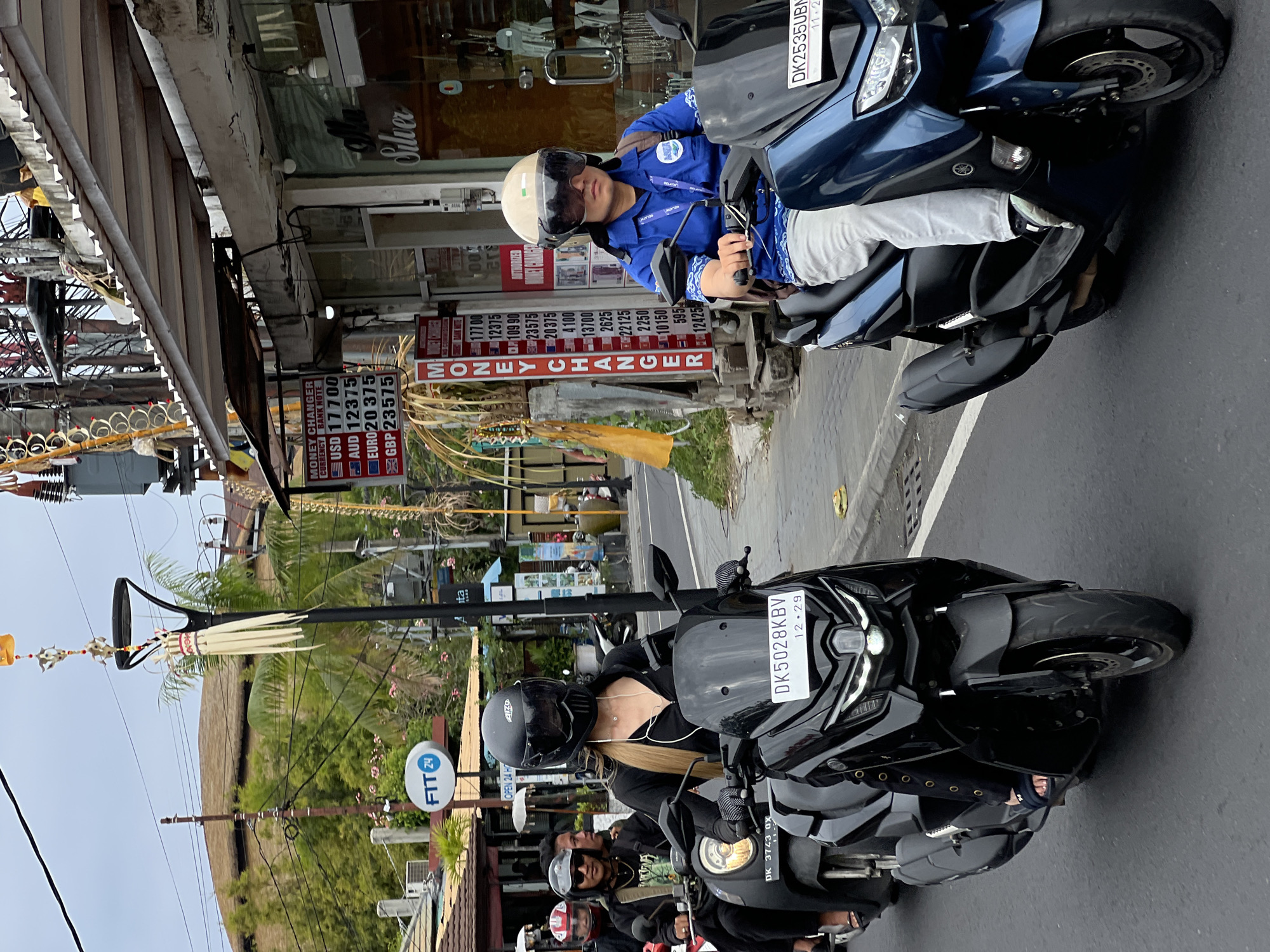 In Denpasar, Indonesia, three individuals are riding scooters on a street. The foremost scooter is black, ridden by a person wearing a black helmet and outfit, with blonde hair visible. To the left, another scooter, also black, carries two people. The driver wears sunglasses and a jacket, while the passenger behind them is partially obscured. The third scooter, blue, is ridden by a person in a blue shirt with a logo, light-colored pants, and a light-colored helmet with a dark visor. All individuals are wearing helmets. In the background, a money changer sign displays currency exchange rates. The street is paved, and a sidewalk is visible to the left with buildings lining the street. Trees and power lines are present in the background. The sky is overcast.