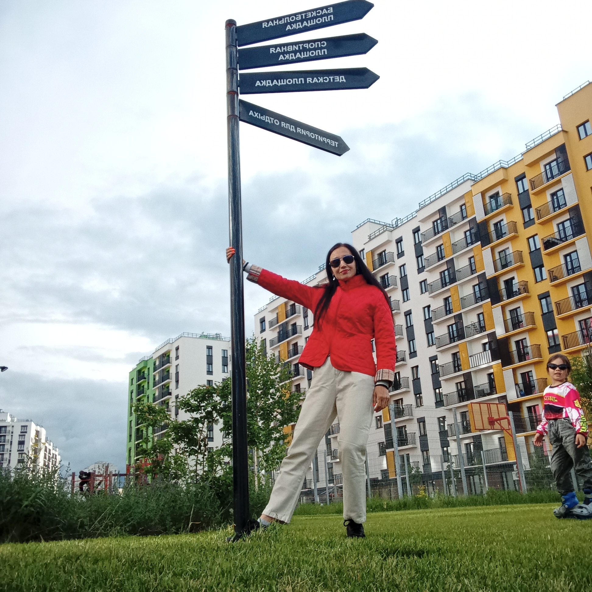 An outdoor, eye-level photograph captures a woman and a child posing near a signpost in a modern residential area in Pervomaysky, Russia. The sky is overcast with gray clouds, indicating a cloudy daytime setting without direct sunlight.
In the foreground and center, an adult woman stands beside a tall, dark signpost, holding it with her right hand. She wears a bright red jacket, light-colored pants, and dark sunglasses, with long dark hair. She has a slight smile, looking directly at the camera. Her fingernails are painted red. The signpost features four dark, arrow-shaped signs with white, mirrored, and largely unreadable text, suggesting a decorative or artistic purpose rather than functional directions. The visible text appears to be in a Cyrillic-based script, but its reversed orientation makes it difficult to decipher.
To the right and slightly behind the woman, a child is visible, also wearing dark sunglasses and a colorful long-sleeved shirt with gray pants, possibly on rollerblades or similar footwear. The child looks towards the camera.
The background features several multi-story residential buildings with balconies, characterized by a contemporary architectural style using a palette of white, yellow, and green facades. Green grass covers the foreground and mid-ground, and some trees and bushes are visible. A fence and a basketball hoop can be seen in the mid-ground, indicating a recreational space within the residential complex. The overall impression is that of a newly developed or well-maintained urban environment.