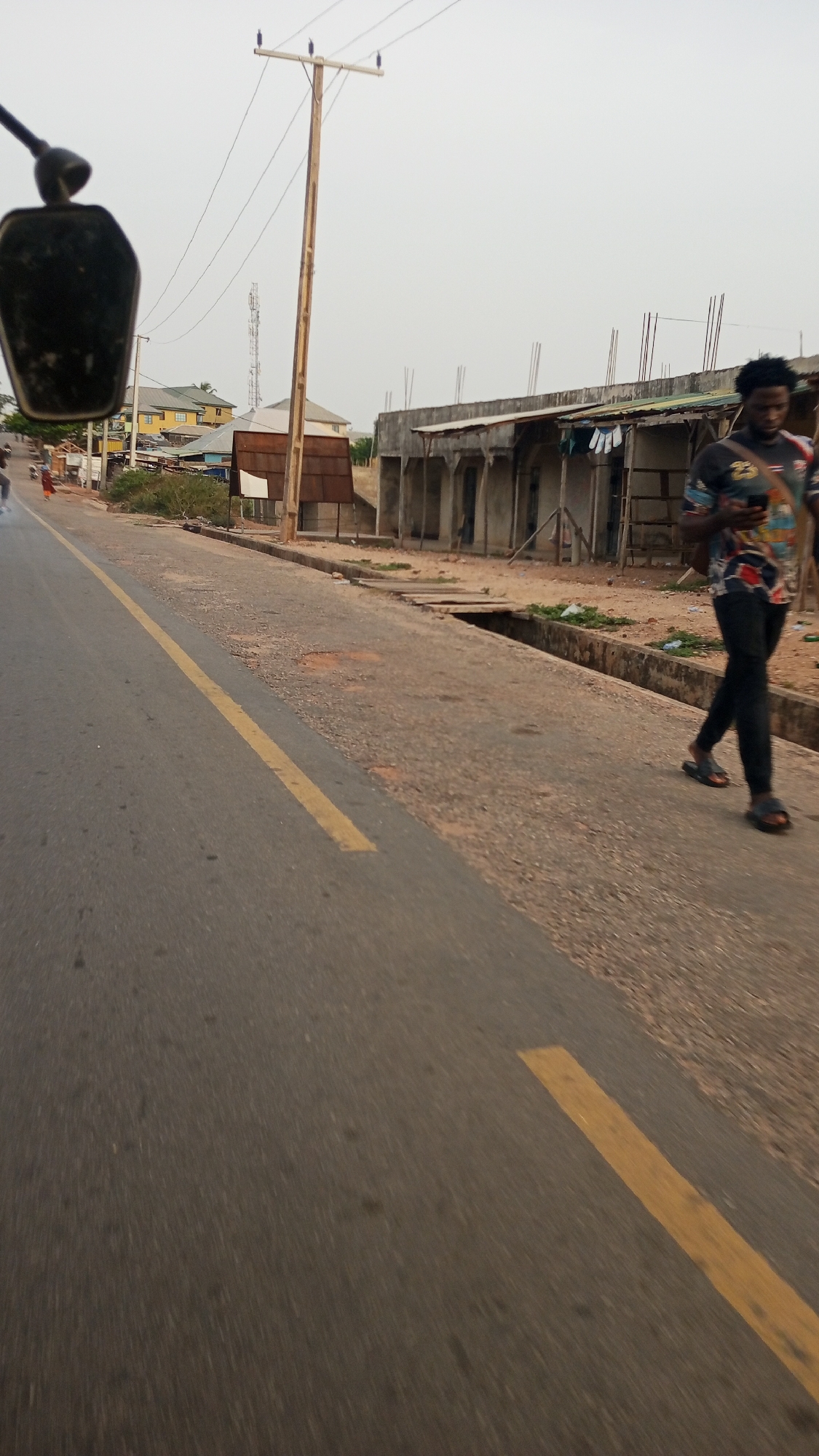 The image depicts a street scene in Zango-ndaji, Nigeria. A man walks on the side of the road near an unfinished building with exposed rebar. The road has a yellow line marking its edge, and there is an open drain alongside the street. A utility pole stands near the building. Other buildings can be seen in the background. A portion of a vehicle's side mirror is visible in the top left corner of the frame.