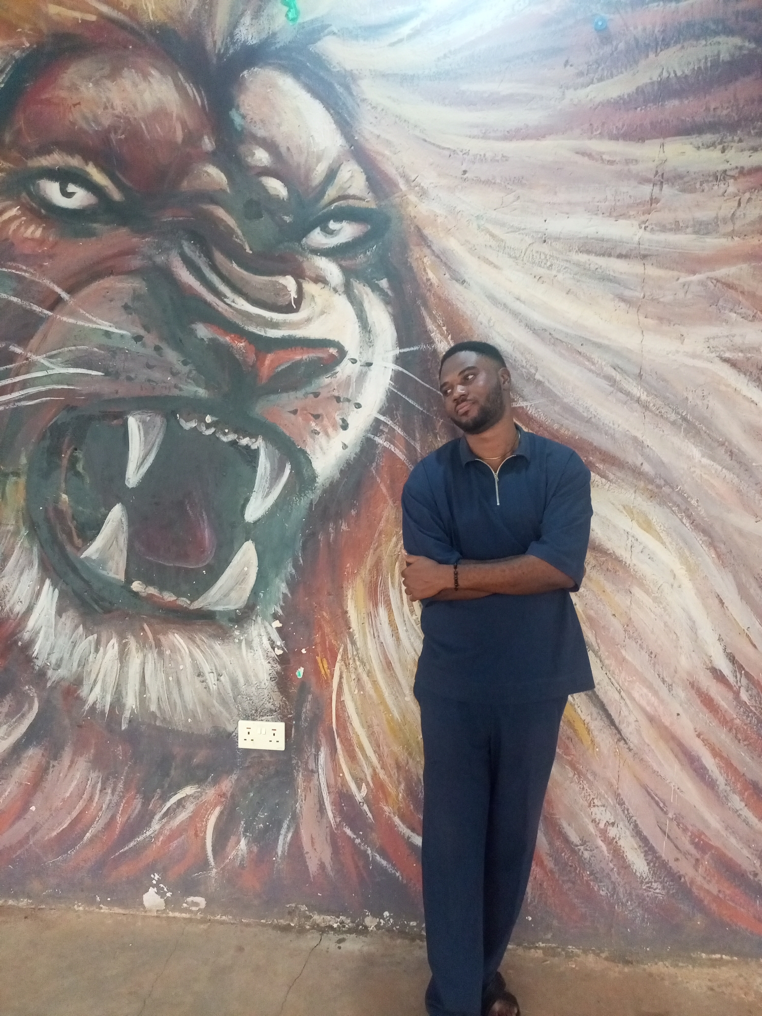 A man in a dark blue two-piece outfit stands with his arms crossed in front of a wall mural of a roaring lion. The mural is predominantly brown and red, and an electrical outlet is visible on the wall near the mural. The man is dark-skinned with a short haircut and a small chain around his neck. The setting is identified as Jos, Nigeria.