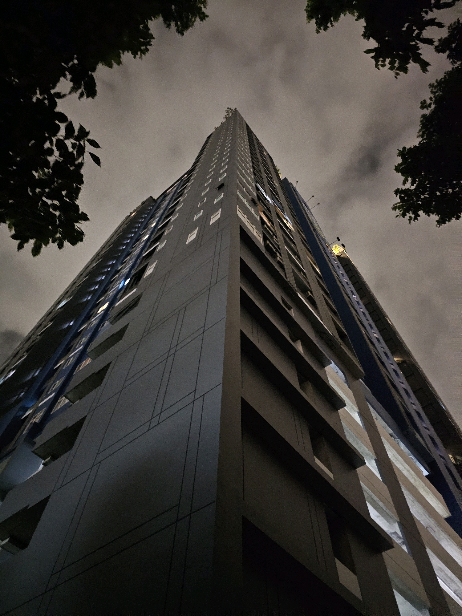 The image captures a low-angle, full-frame view of a tall, modern apartment building at night in Bangkok, Thailand. The building, predominantly gray with blue accents and numerous lit windows, rises steeply towards a dark, cloudy sky. The perspective emphasizes the building's height and scale, with the top portions of two trees silhouetted against the sky in the upper left and upper right corners of the frame. The architecture features distinct horizontal and vertical lines, with sections of the facade appearing to be cantilevered or recessed. The overall atmosphere is moody and somewhat dramatic due to the nighttime lighting and overcast sky. No people are visible in the image.