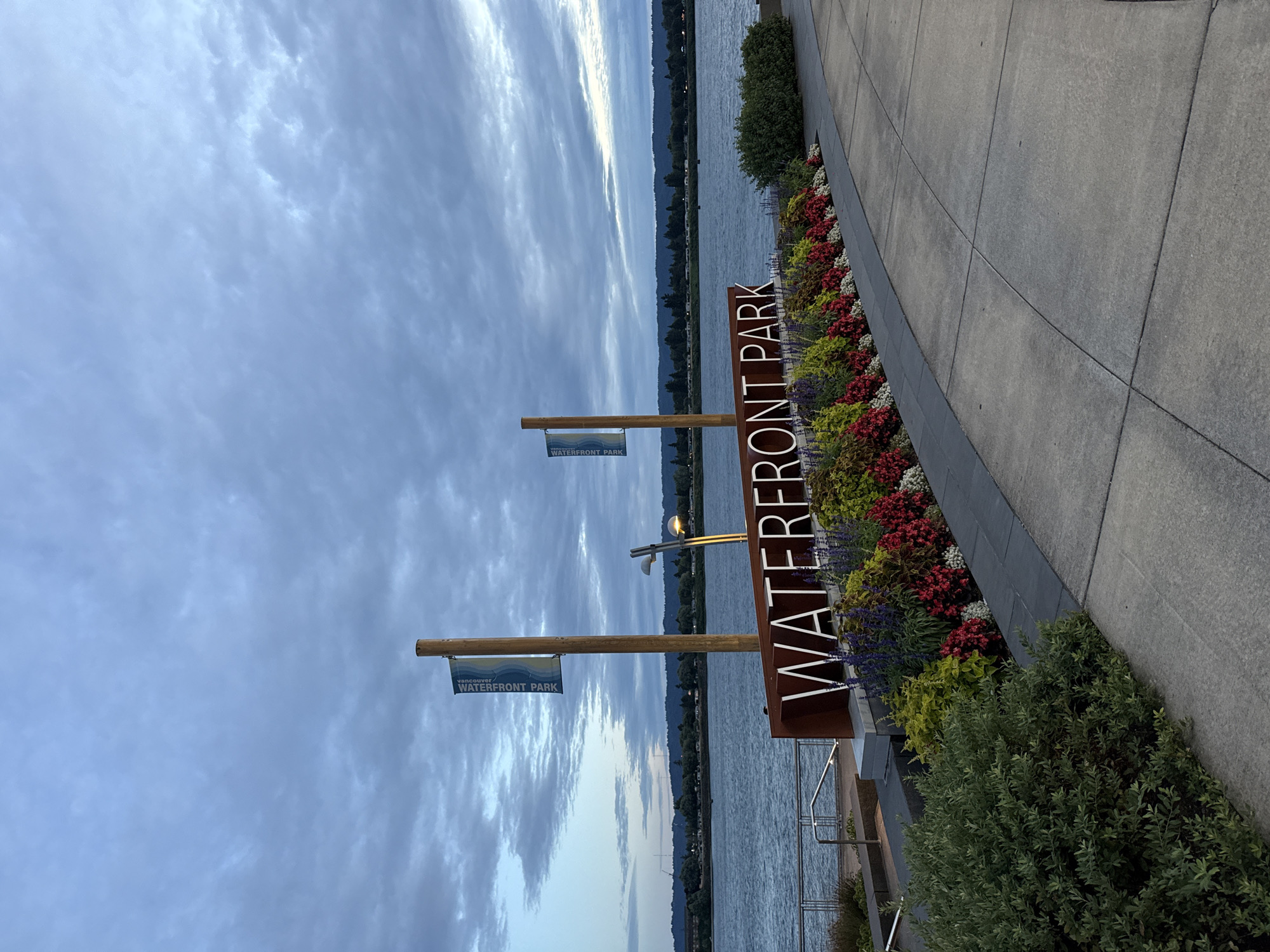 The image shows a sign that says "WATERFRONT PARK" in large white letters, situated in front of a colorful flower bed with red, yellow, and purple flowers. Behind the sign, there is a body of water, likely a river or bay, with a distant tree-lined shore. The sky above is cloudy and appears to be either early morning or late evening. A wooden pole with a blue banner reading "WATERFRONT PARK" stands to the left of the sign. The paved walkway in the foreground is made of large concrete slabs. The scene is located at Waterfront Park in Vancouver, Washington, United States of America.
