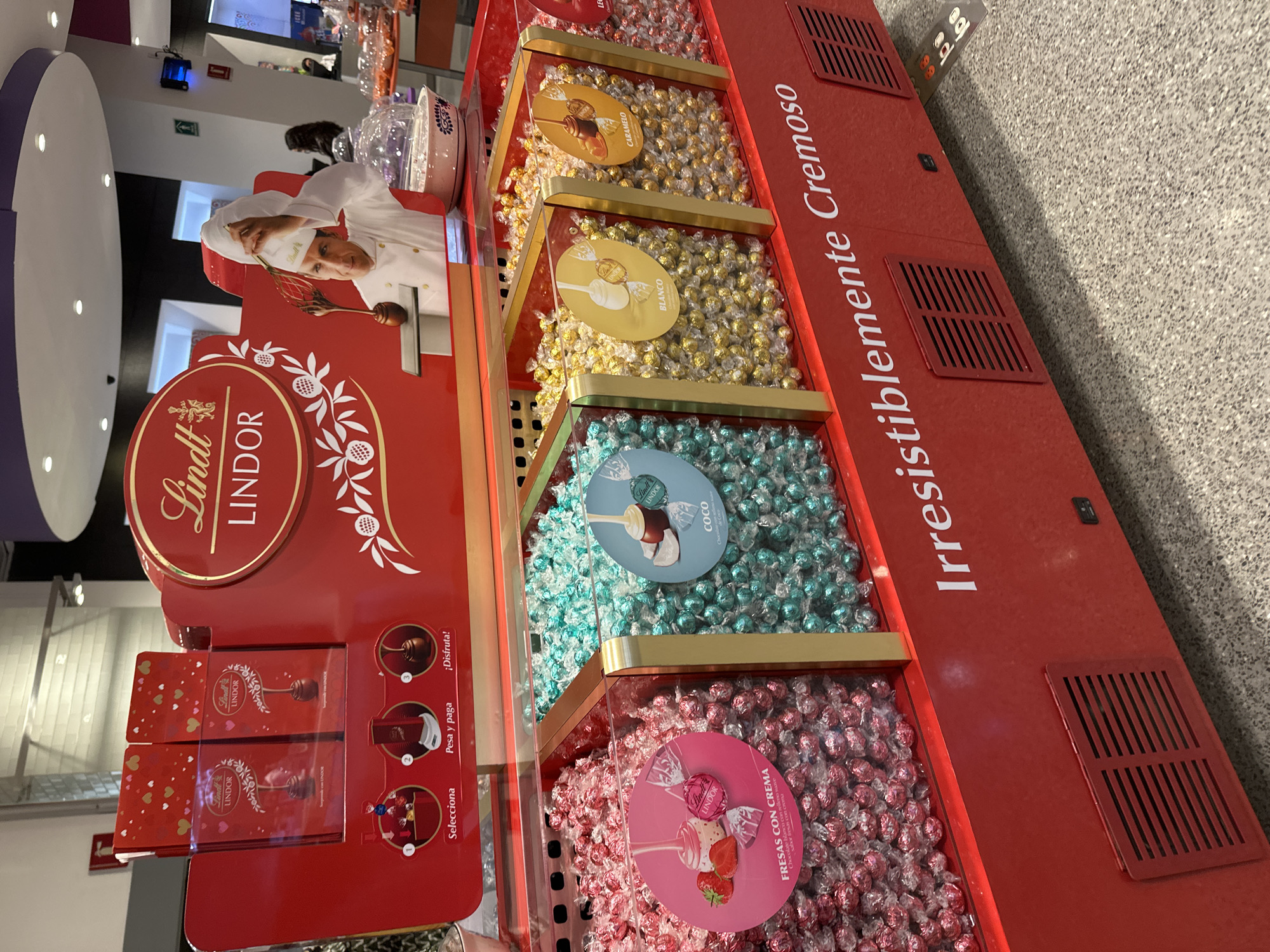 The image shows a Lindt Lindor chocolate display stand, located in what appears to be a shopping center in Puebla, Mexico, due to the Spanish text on the display. The stand is red and features multiple tiers of loose Lindor chocolates in various flavors, including caramel, white chocolate, coconut, and strawberry cream. Each tier is labeled with a round flavor sign. The stand also includes a graphic of a chef stirring chocolate and instructions for selecting, weighing, and enjoying the chocolates. The text "Irresistiblemente Cremoso" is displayed prominently along the side of the stand. The flooring of the space where the stand is located appears to be grey tile.