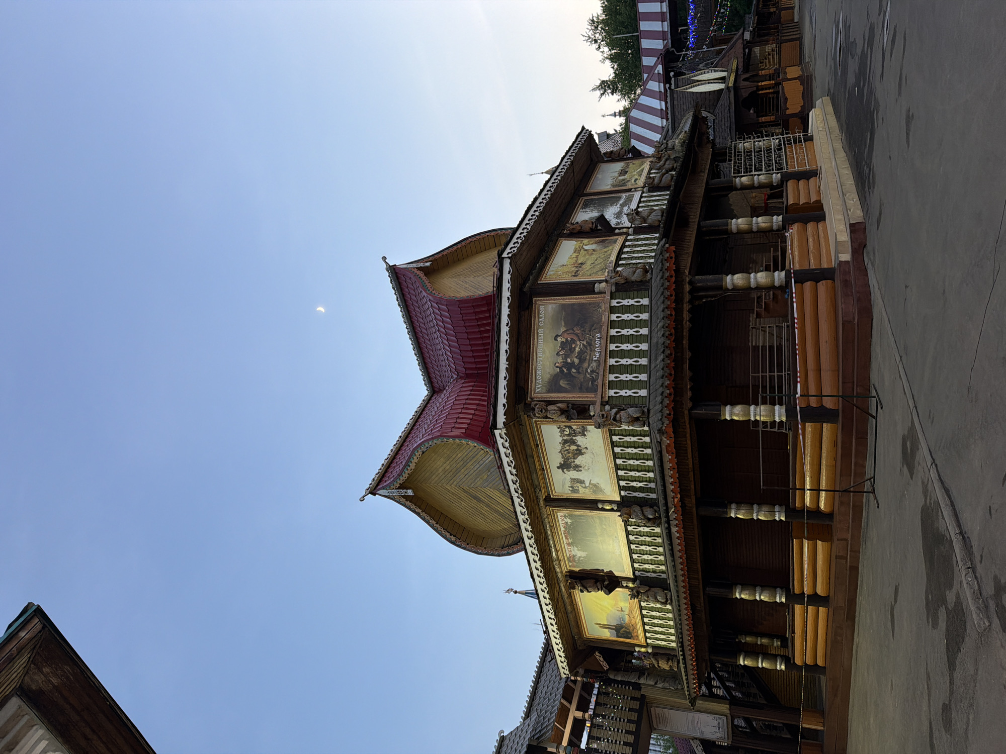 The image captures a view of a traditional Russian wooden building located in Moscow, Russia. The architectural style is characterized by intricate wooden carvings and painted details on the exterior. The building features a red roof and a balcony with framed paintings displayed along the facade. The structure stands against a clear blue sky, with a hint of a lunar phase visible. The surrounding environment suggests an urban setting, likely within a historical or cultural area of Moscow.