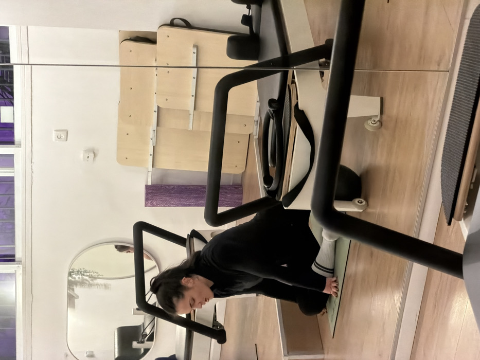 A woman in black athletic wear and striped socks practices Pilates on a reformer machine, which is reflected in a wall mirror. The room is white with a hardwood floor. Additional equipment includes wall-mounted bars and a purple foam roller. The woman bends forward, engaging with the apparatus.