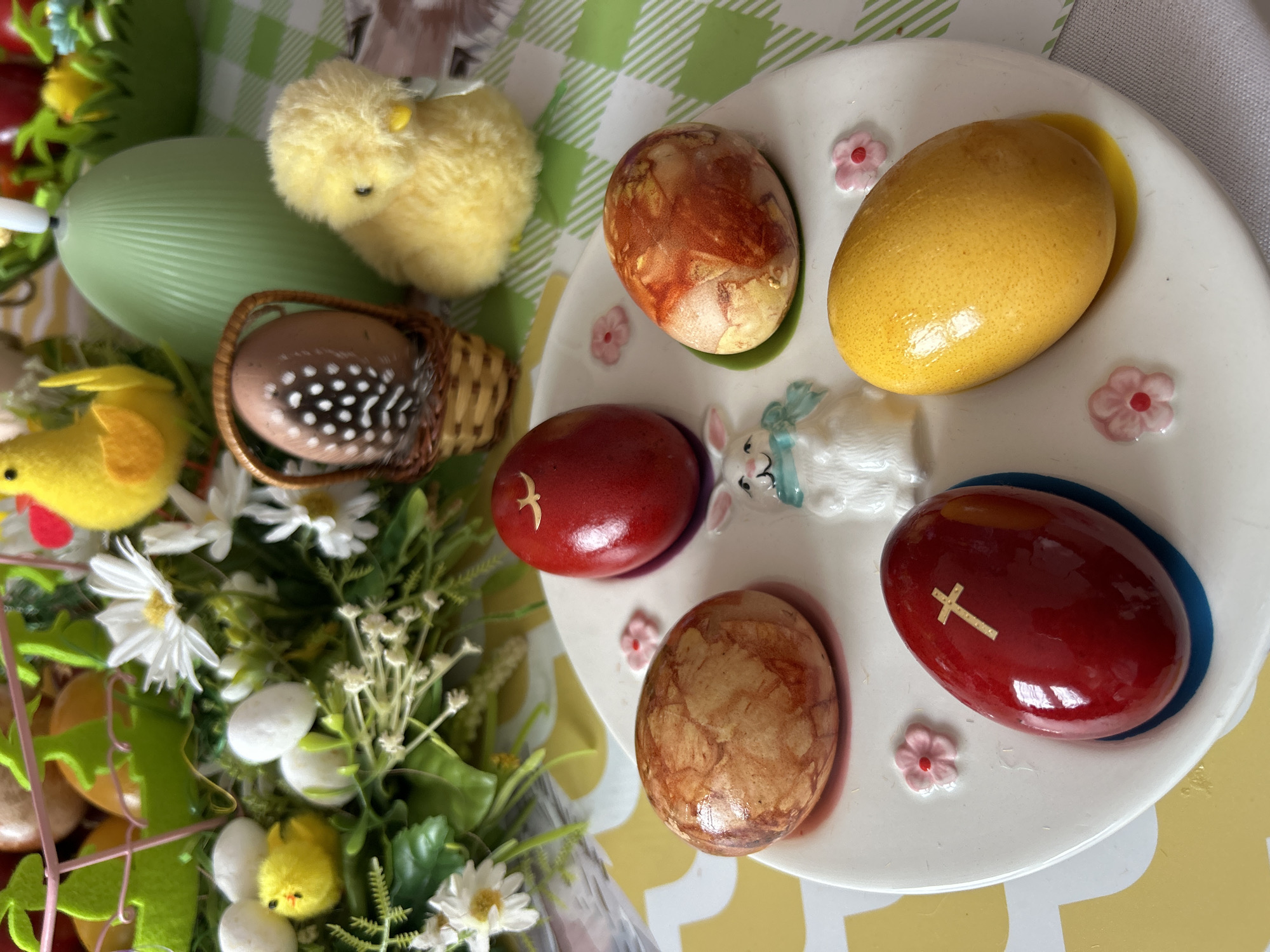 The image shows an Easter scene. On a white plate with decorative pink flowers, there are five decorated eggs. The eggs are colored in shades of yellow, brown, and red. One red egg has a gold-colored cross on its surface, and another has a gold-colored bird. Between the eggs, there is a small porcelain bunny. Next to the plate, there is a woven basket containing a speckled egg and a feather. Additionally, there are yellow chick decorations, a green decorative egg, and a bouquet of flowers, including white daisies. The background appears to be a checkered tablecloth. The scene does not show anything that indicates the location in Plovdiv, Bulgaria.