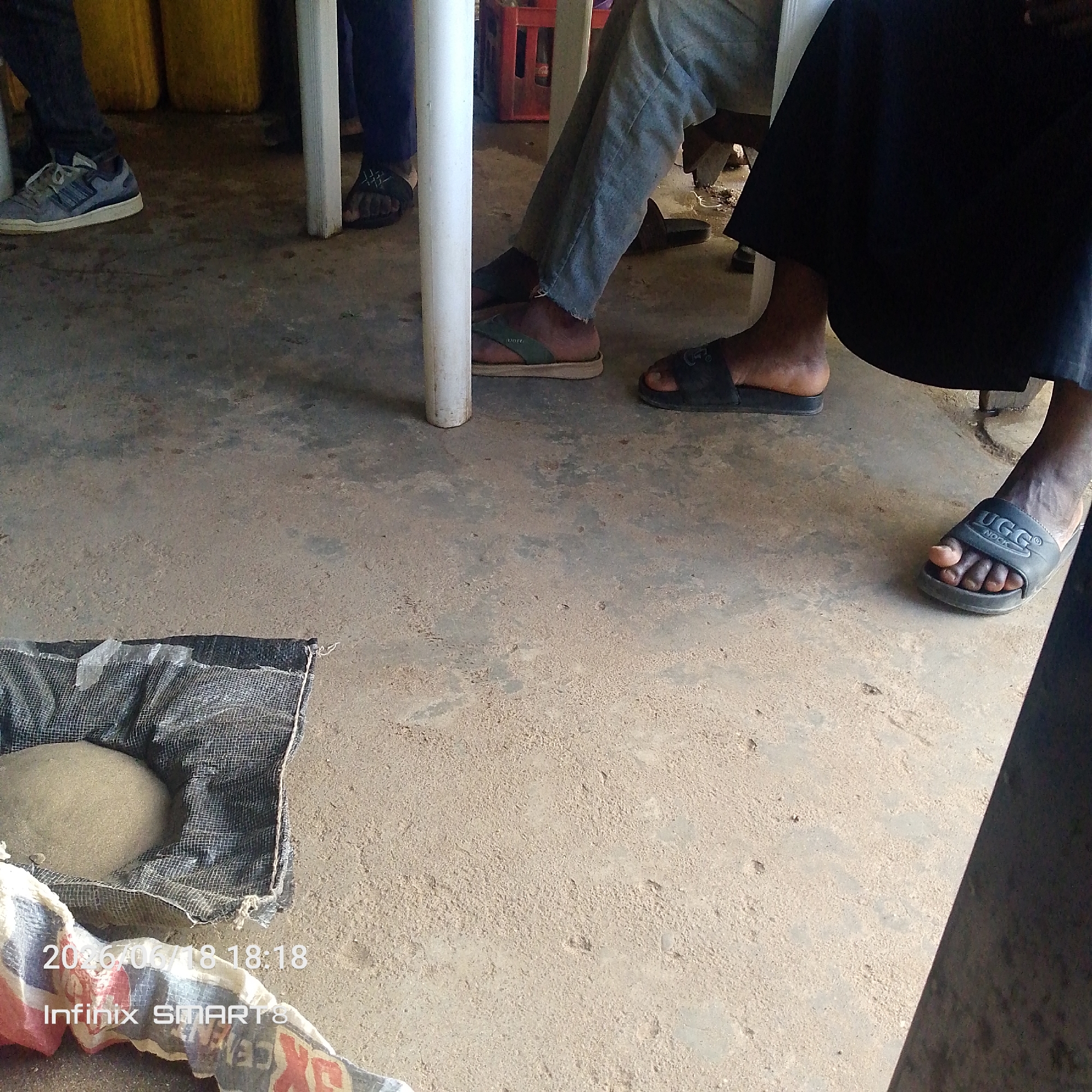 The image shows the lower bodies of at least three people standing or sitting around a concrete floor. The scene appears to be outdoors or in a very open indoor space, with a sandy or dusty floor. In the foreground, there's a partially visible object wrapped in a black mesh bag, and a large, light-colored, rounded object inside it, possibly a ball of dough or earth. There's also a piece of fabric with colorful patterns, possibly part of a bag or cloth, in the bottom left corner.
One person is wearing grey pants and green and beige sandals. Another person is wearing dark pants and black flip-flops with a white logo. A third person is wearing dark pants and black slides with a white logo that appears to say "UGG NOOK". A fourth person's feet are visible on the left, wearing light blue and white sneakers. In the background, there are large yellow barrels.
The image has a timestamp in the bottom left corner that reads "2023/06/18 18:18", suggesting it was taken in the evening. There is also text that reads "Infinix SMARTS". The lighting suggests it might be daytime, possibly late afternoon, with some shadows cast on the floor. The overall mood is casual and perhaps utilitarian, given the presence of the materials in the foreground.