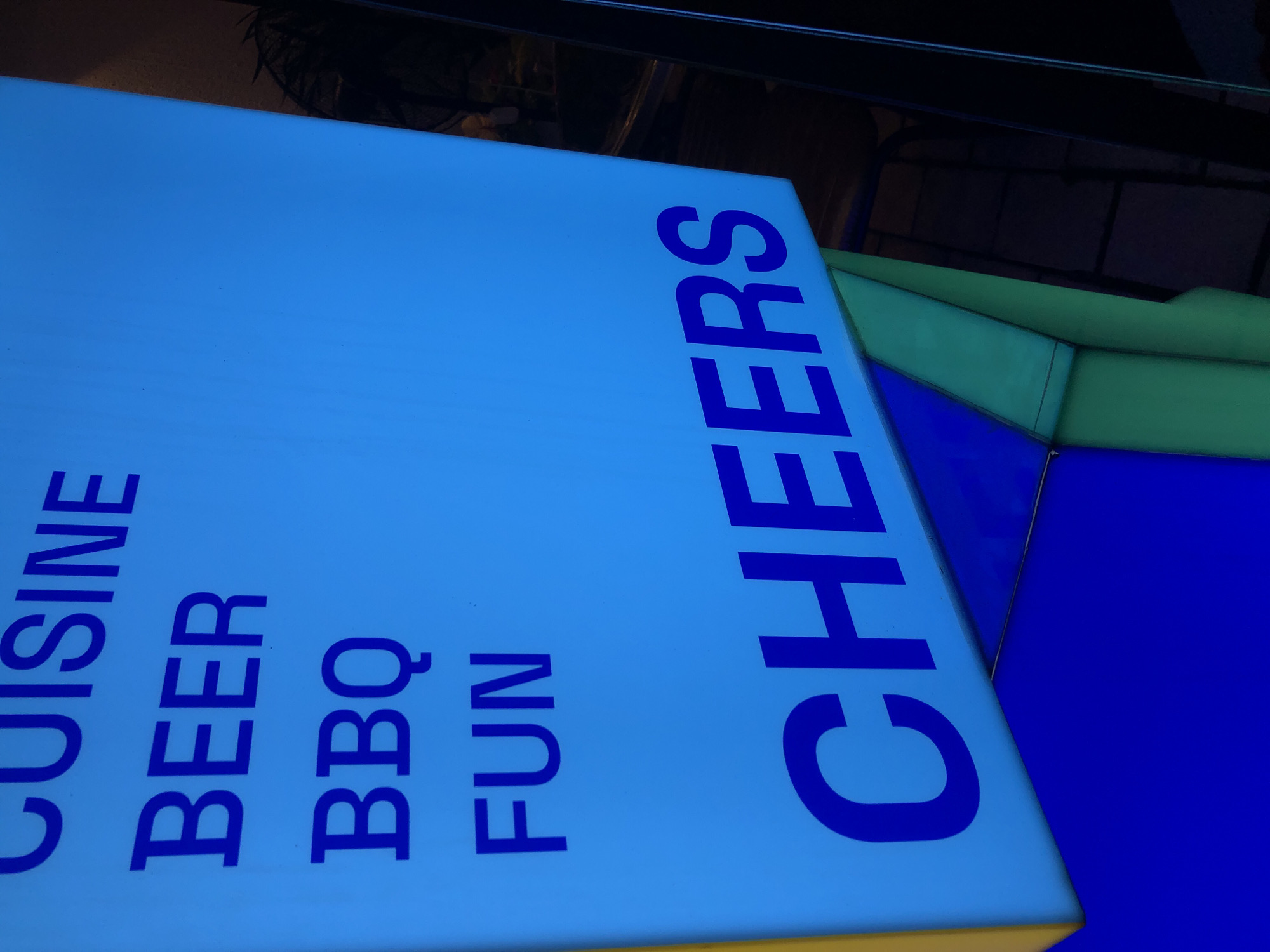 The image shows a close-up of a brightly lit blue sign with blue text. The text includes the words "CUISINE", "BEER", "BBQ", "FUN", and "CHEERS". The sign is angled, with the top left corner appearing closer to the viewer. The background is dark and out of focus, with some hints of greenery and what appears to be a metal structure. The sign is likely an advertisement for a restaurant or bar. No specific location in Da Nang, Vietnam is identifiable from the image.