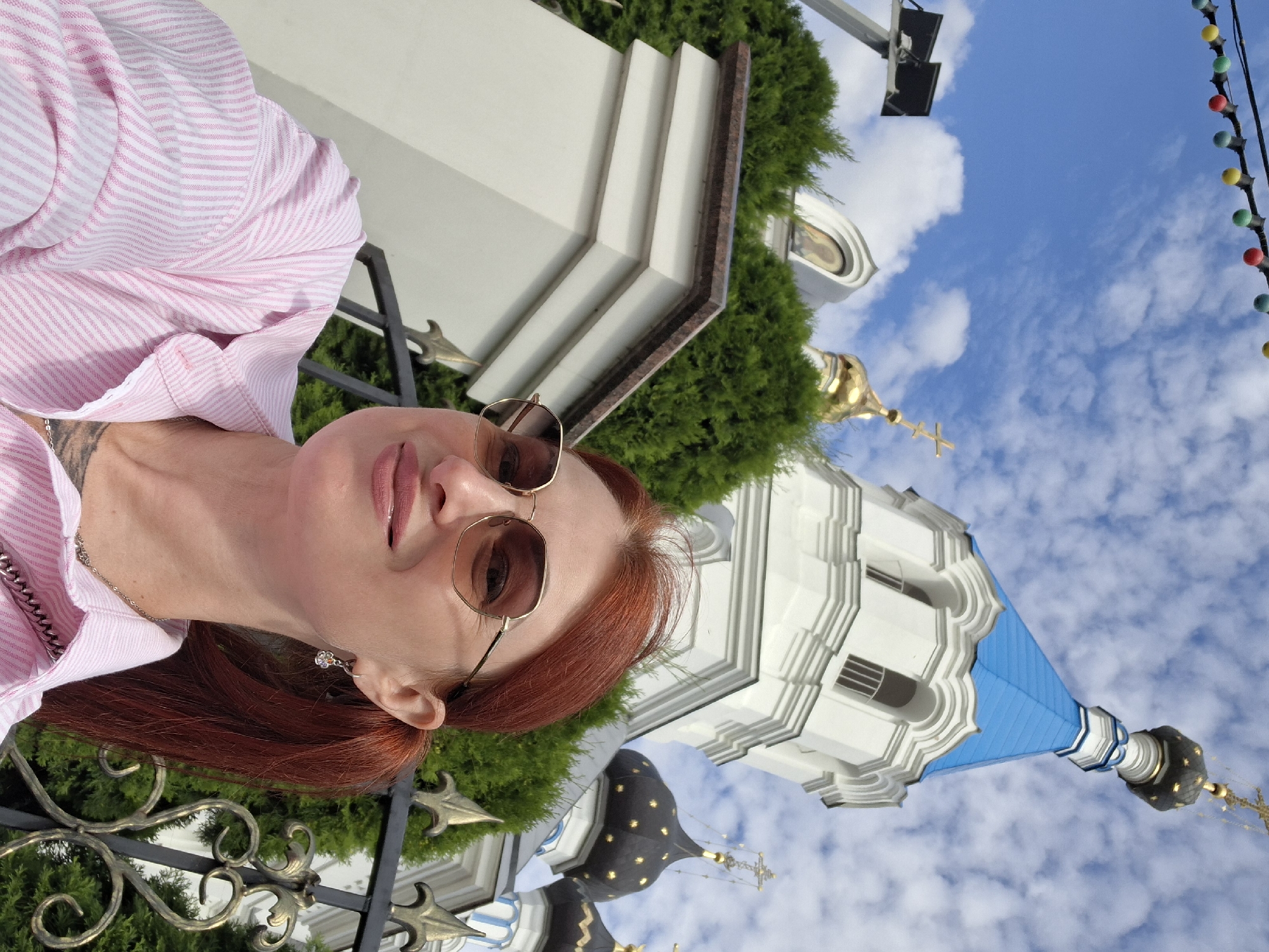 A woman with red hair and sunglasses smiles at the camera in front of a white church with a blue roof and gold domes. She is wearing a pink and white striped shirt. The church appears to be a Russian Orthodox church and the scene is likely the Holy Transfiguration Church in Pastavy, Belarus. The sky is blue with white clouds, and there are some trees and a black metal fence in the foreground. There is also a string of colorful lights visible at the top of the image.