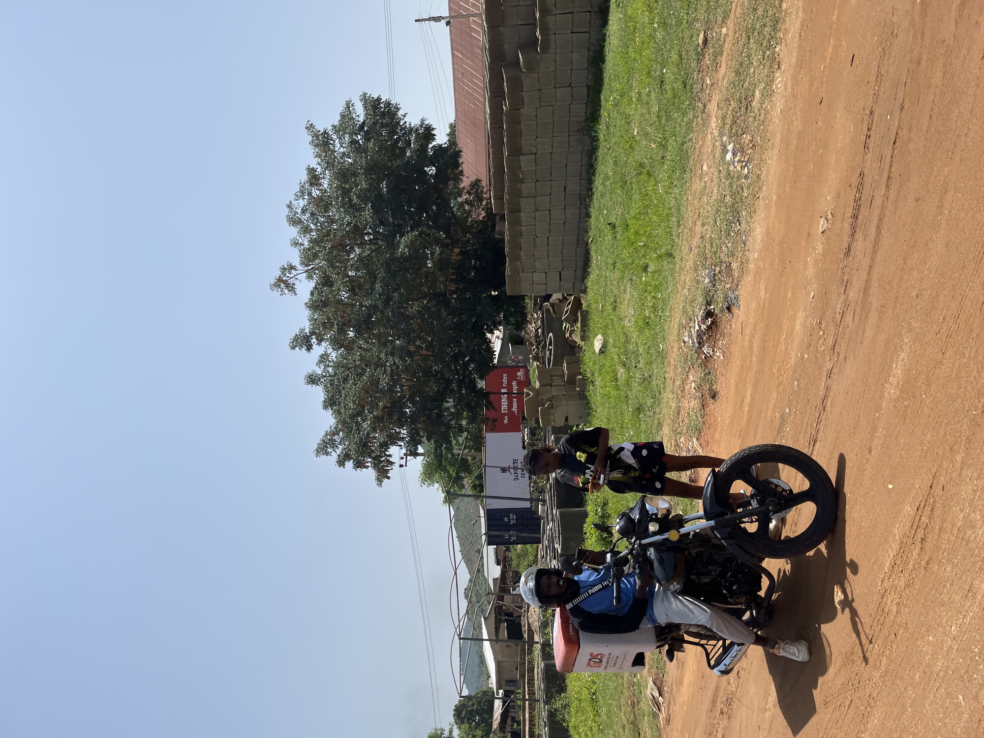 In Gwong, Nigeria, a man wearing a helmet and a blue long-sleeved shirt, seated on a black motorcycle, appears to be pausing on a dirt road. The motorcycle has a white and red delivery box attached to the rear. Next to him stands a woman wearing a black t-shirt and patterned shorts. She is holding a phone. The environment suggests an outdoor setting with a partially built structure, stacks of concrete blocks, a large tree, and a clear blue sky. In the background, a mountainous landscape is visible.