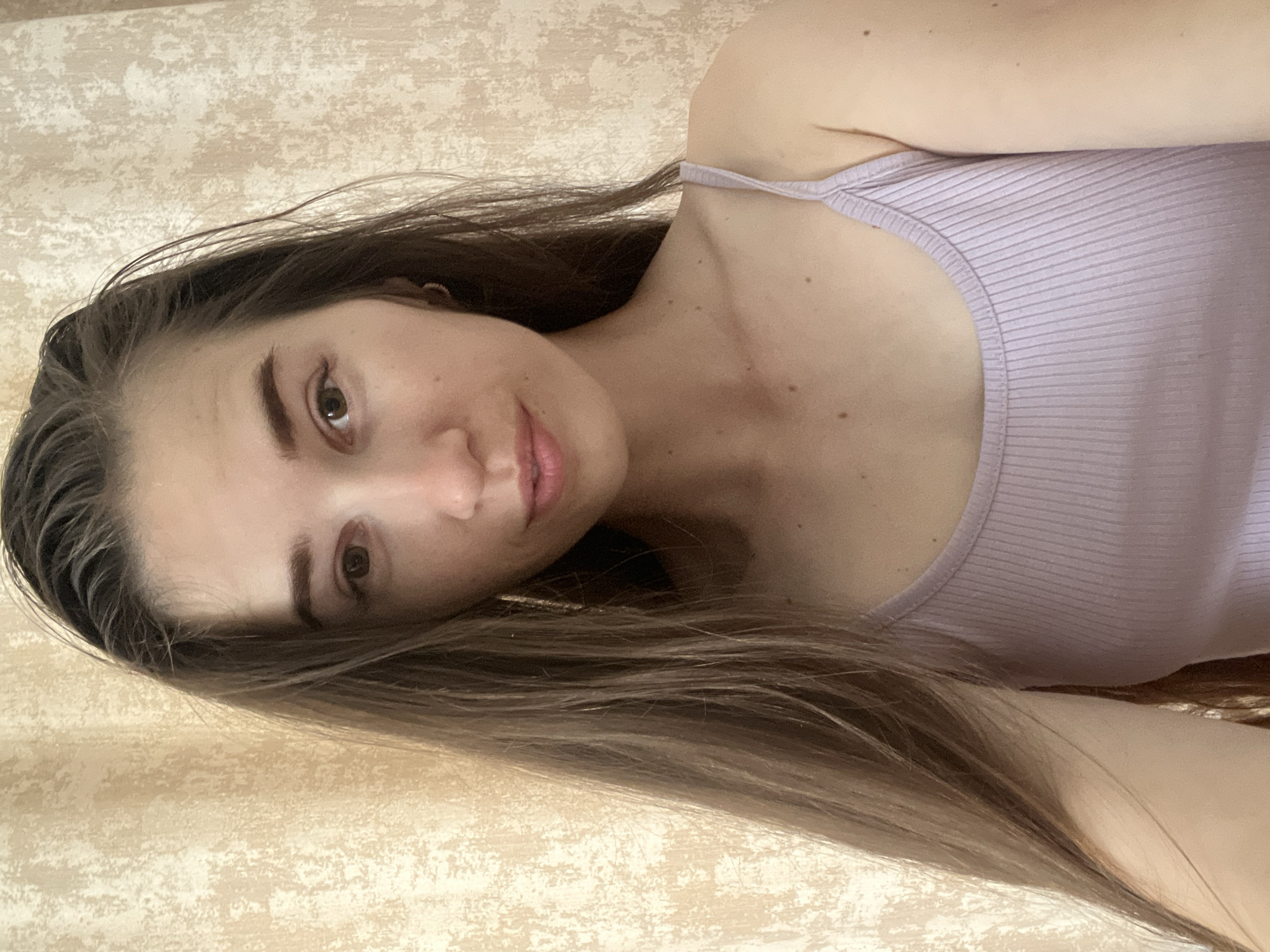 A young woman is pictured in a close-up selfie against a beige, textured wallpaper. She has long, dark brown hair that falls past her shoulders. Her gaze is directed towards the camera, and she has light brown eyes and light pink lips. She is wearing a lavender ribbed tank top with thin spaghetti straps. A small, gold hoop earring is visible in her left ear. The lighting suggests an indoor setting.