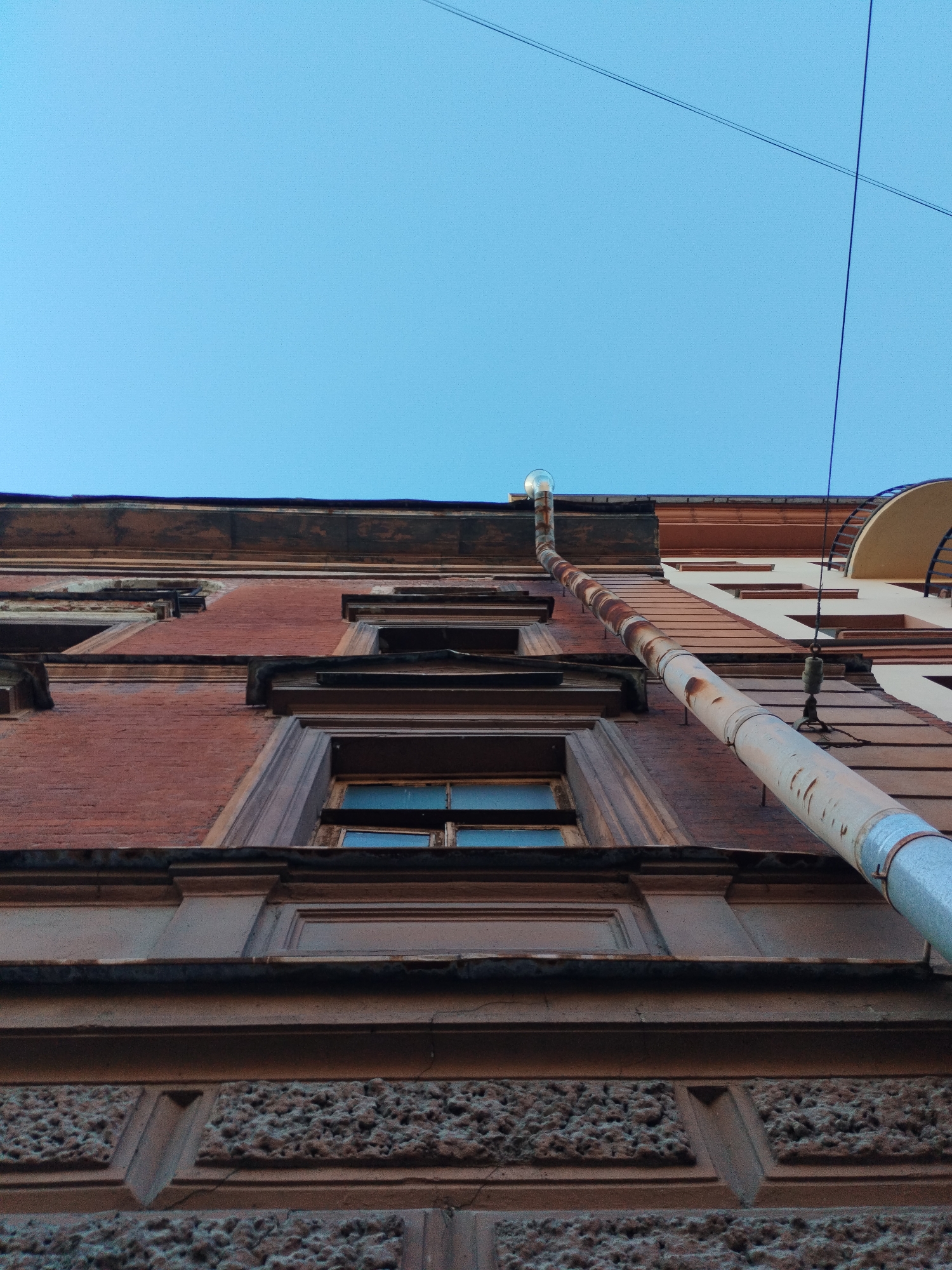 A low-angle, upward shot shows the facade of a multi-story building in Saint Petersburg, Russia, against a clear blue sky. The building's brickwork is a muted red, with decorative brown trim and ornamentation visible around windows and along horizontal ledges. A prominent, rusty metal downspout runs diagonally across the frame, appearing to be attached to the building at various points. Above the downspout, a single overhead wire is visible against the sky. The architectural style suggests an older, possibly historic, building, common in the city's many residential areas. The image captures the verticality of the structure and the contrast between the weathered facade and the bright sky.
