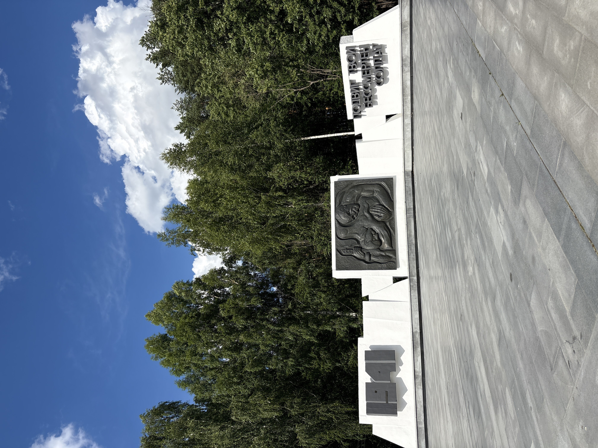 This media file depicts a war memorial in Chaikovsky, Russia. The memorial consists of a white, tiered structure with inscribed text and a bas-relief sculpture. The text, written in Cyrillic, reads "ПОДВИГ ТВОЙ БЕССМЕРТЕН, СОЛДАТ" which translates to "Your feat is immortal, Soldier." Below this inscription, the numbers "1941" are prominently displayed, likely referencing the year 1941, a significant year during World War II. The bas-relief appears to depict human figures in a somber or reflective manner.
The memorial is situated outdoors, with a bright blue sky and lush green trees forming the backdrop. Several white, fluffy clouds are visible in the sky, suggesting it is a clear day, likely midday or afternoon due to the strong sunlight casting shadows. The ground in front of the memorial is paved with grey stone tiles. The overall atmosphere evoked by the imagery is one of solemnity and remembrance.