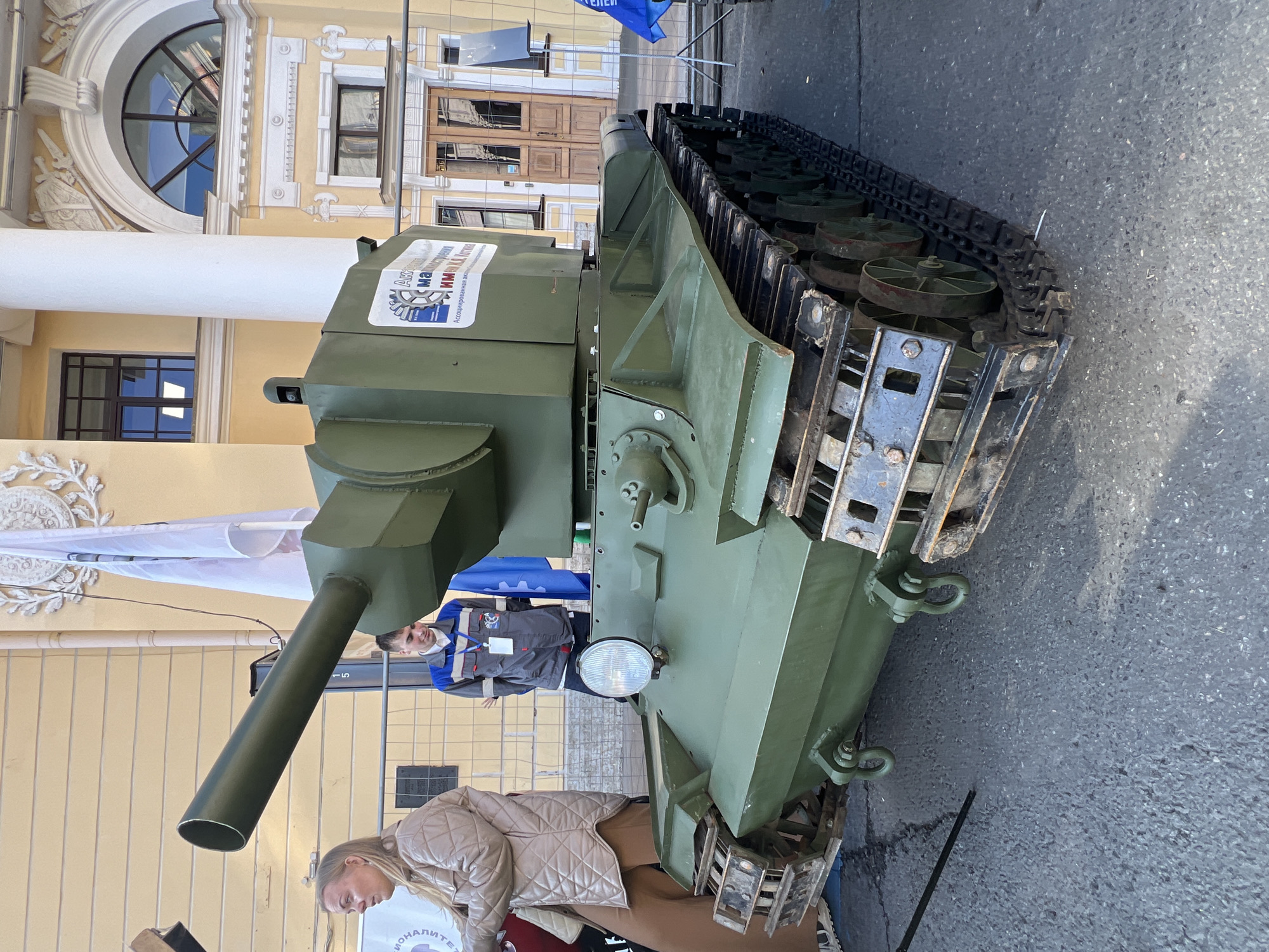 A green model of a military tank is parked on the asphalt in front of a light-yellow building. The tank model includes a long, cylindrical gun barrel, tracks, and several people are standing nearby. A woman with blond hair is visible near the tank's front, while a man in a grey uniform is partially visible behind the tank's turret. A white and blue banner hangs behind the tank. The building in the background has arched windows and classical architectural details, a white column is also seen.
Based on the architecture, it is likely that the location is in Saint Petersburg, Russia. The architectural style and the building color match the historical buildings in that city.
