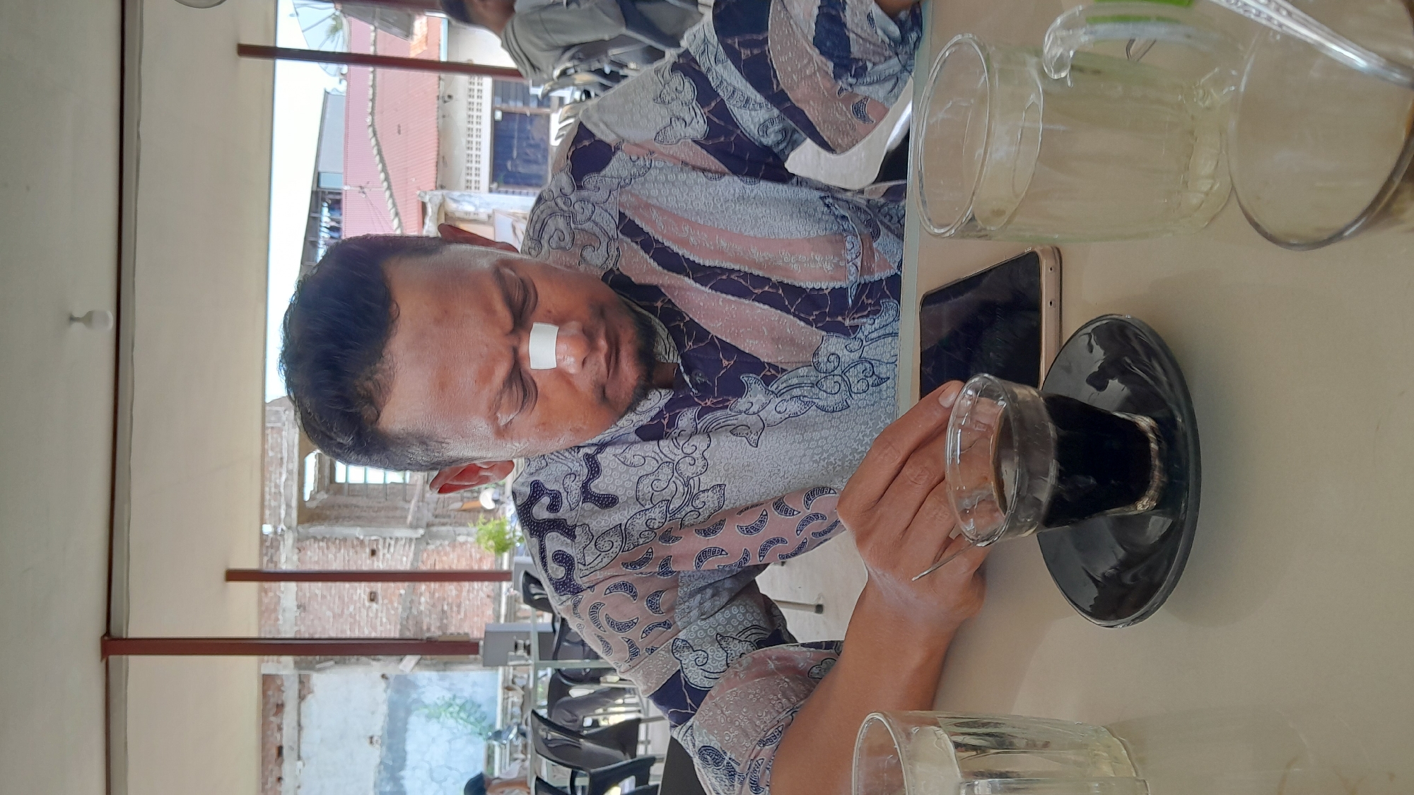 The image captures a scene within an open-air structure in Lhokseumawe, Indonesia. An adult male with a bandage on his nose sits at a table, wearing a patterned shirt. He appears to be looking at a dark-colored beverage in a glass, resting on a shiny black saucer. A smartphone lies near the glass. There are two empty glass pitchers on the table. The background shows the urban environment, including what appears to be a building under construction or renovation. There are chairs visible, suggesting a cafe or restaurant setting.
