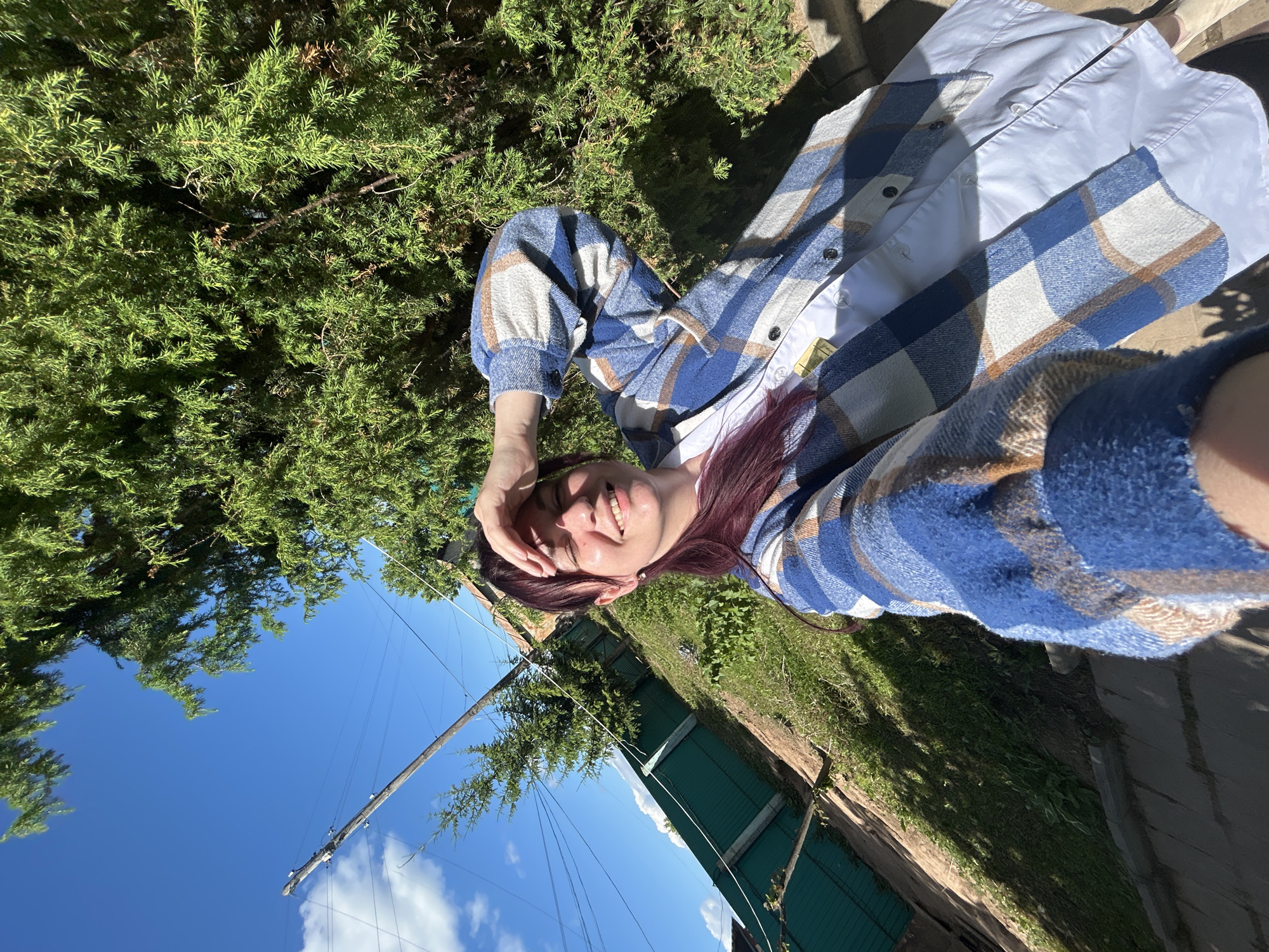 A young woman with dark hair, wearing a blue and white plaid jacket over a white shirt, is taking a selfie outdoors. She is shielding her eyes from the sun with her hand and smiling. The background features a lush green tree, a blue sky with some clouds, a green fence, a wooden utility pole with wires, and a grassy slope. The ground in the foreground appears to be a pathway made of concrete or stone. The image captures a bright, sunny day in what is identified as the city of Fokino, Russia.