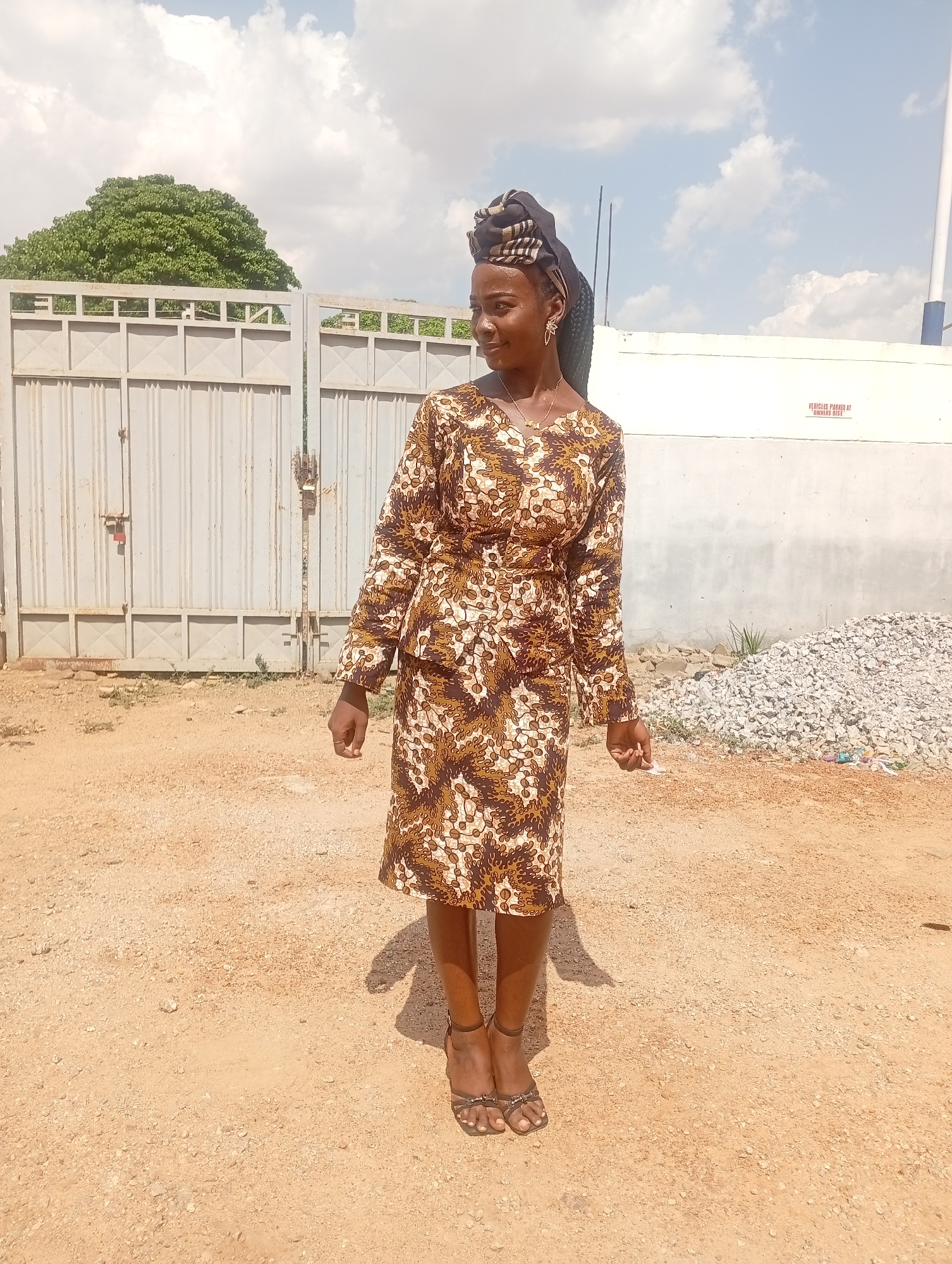 A young woman stands in a dusty, possibly unpaved area, seemingly in Sabon Tasha, Nigeria, based on the environmental conditions and the style of dress. She is wearing a brown and white patterned dress with long sleeves and a matching head wrap. Her feet are clad in dark brown sandals. Behind her, there is a light gray metal gate on the left, a white wall with a warning sign on the right, and a pile of rocks. A green tree is visible beyond the gate, and the sky is partly cloudy.