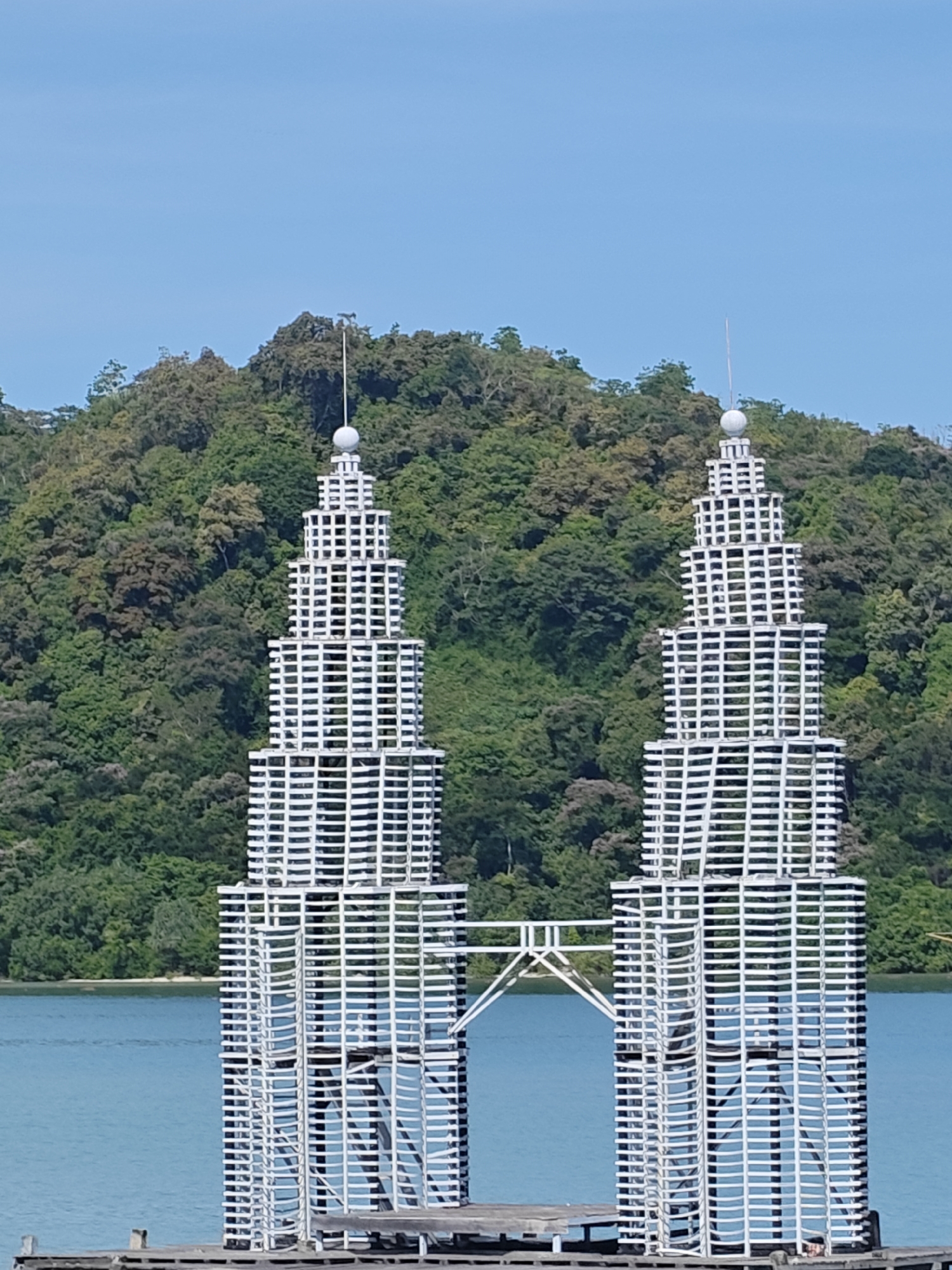 The image depicts a scaled-down model of the Petronas Twin Towers, constructed from white latticework, situated on a platform in a body of water. A bridge-like structure connects the towers. Lush green foliage covers a hillside in the background. The sky is clear and blue. The towers are identified as being located in Layeun, Indonesia.