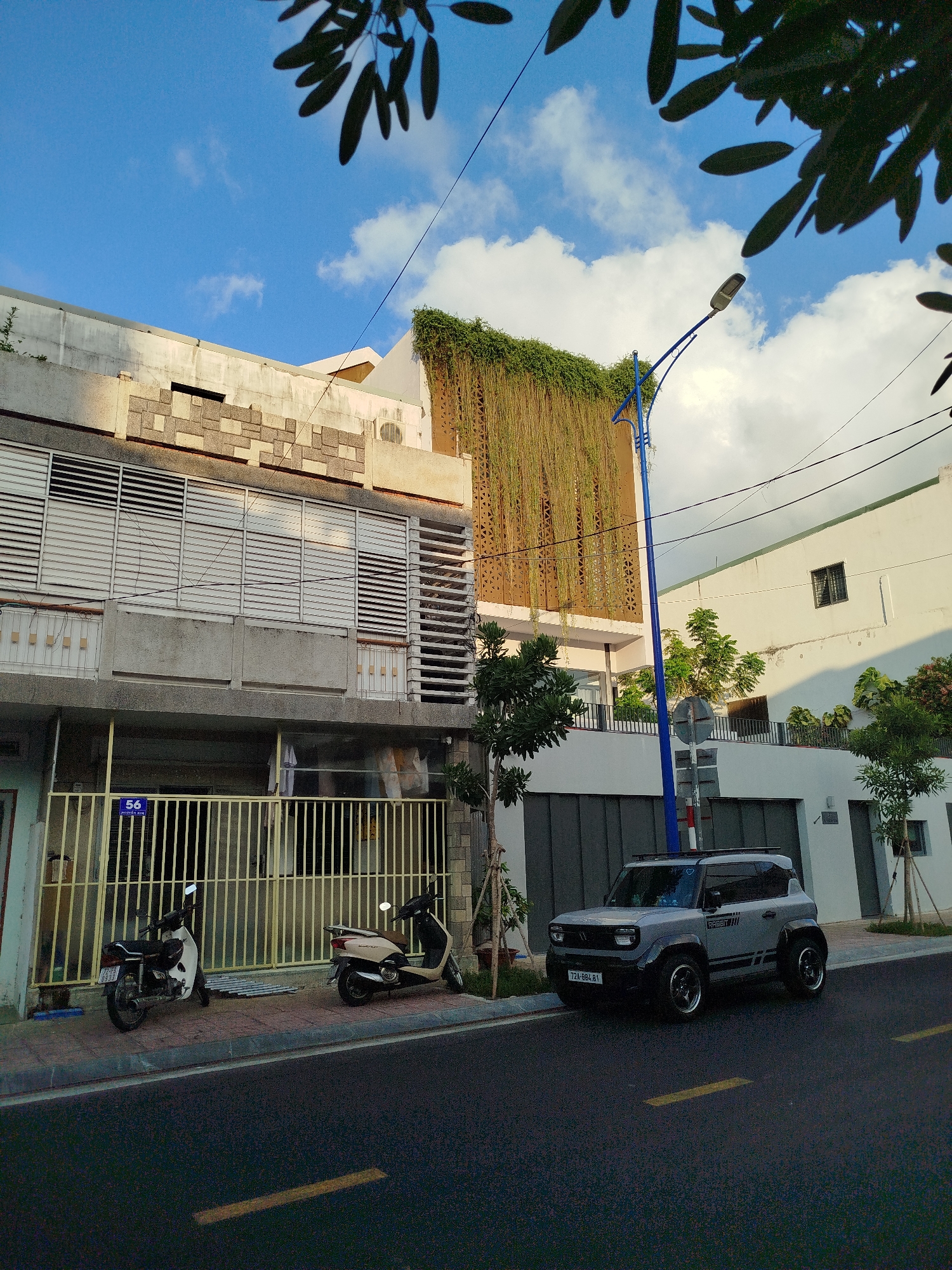 The image shows a street scene in Ho Chi Minh City, Vietnam. A gray electric vehicle is parked on the right side of the street, adjacent to a white building with a modern design. To the left of the EV, there is a two-story building with a concrete facade, featuring horizontal window slats. A green vine-like plant hangs down from the upper part of the building on the right. A couple of motorcycles are parked behind a yellow gate at the front of the concrete building. A blue street lamp stands near the EV. Trees and branches are visible in the background and foreground. The sky is partly cloudy.