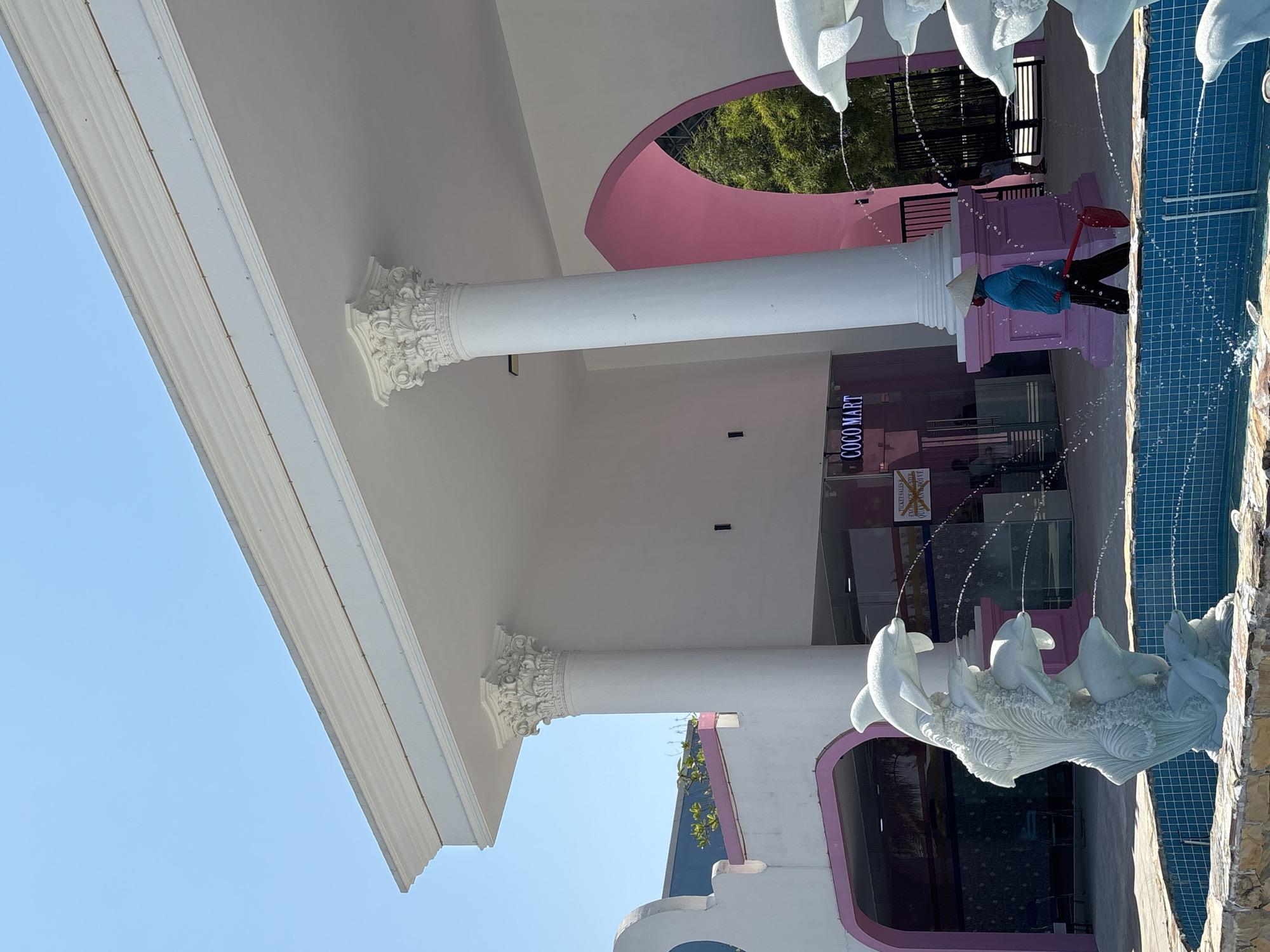 The image shows the exterior of a commercial building, notably the "COCO MART" store in Đà Nẵng, Vietnam. The building has white columns with ornate Corinthian capitals and a white roof. A pink arch detail surrounds the store's entrance. There is a dolphin fountain with water flowing into a blue pool in front of the building. A person wearing a blue shirt, dark pants, and a conical Asian hat is cleaning the area with a red tool near the fountain.