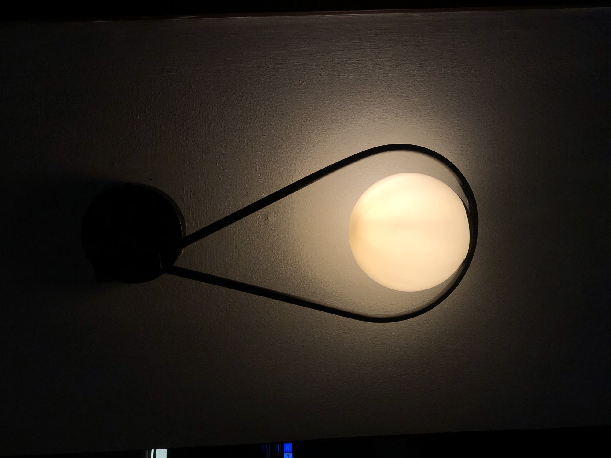The image features a modern wall-mounted light fixture against a textured, light-colored wall. The fixture consists of a spherical, frosted white glass globe that emits a warm, soft yellow-orange light. This globe is encased within a sleek, dark-colored (likely black) metal frame that forms an elongated, somewhat organic loop around the globe, connecting to a dark, circular base attached to the wall.
The light emanating from the fixture illuminates the immediate area of the wall, creating a glowing aura around it, while the surrounding parts of the room are plunged into darkness, indicating a low-light environment or nighttime. The contrast between the brightly lit wall section and the deep shadows is stark. The wall surface shows a subtle, uneven texture, possibly painted plaster or drywall, which becomes more apparent where the light hits it. There are no people or discernible activities taking place. The design of the light fixture suggests a minimalist or contemporary aesthetic. No visible text is present in the image.
