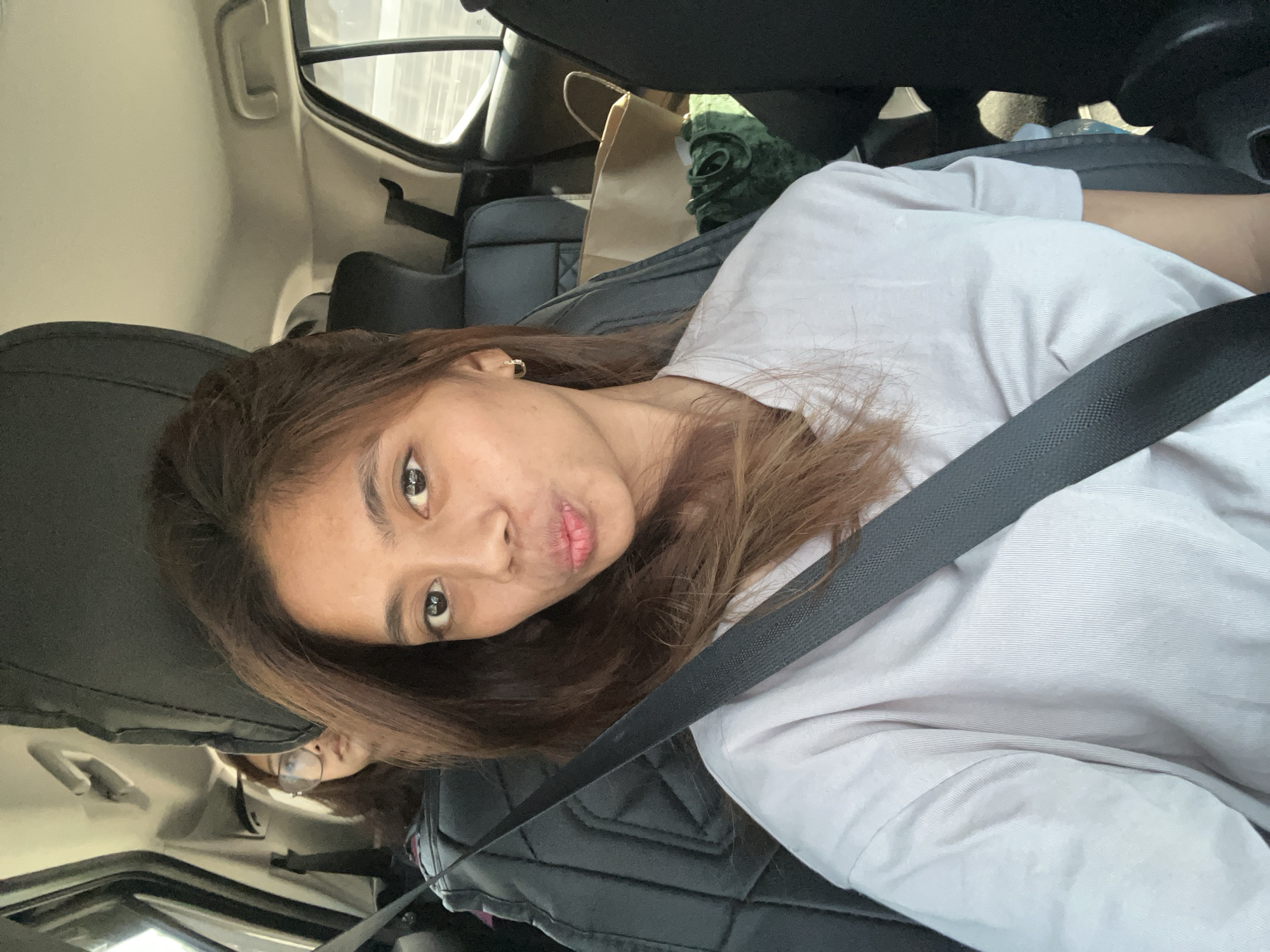 A selfie-style photograph captures two female occupants inside a modern vehicle in Davao City, Philippines, during daylight hours. The primary subject, a woman with medium brown, shoulder-length hair, occupies the front passenger seat. She wears a light grey, short-sleeved t-shirt with subtle vertical ribbing and is secured by a black seatbelt diagonally across her chest and lap. Her eyes are dark, looking directly at the camera, and her lips are pursed in a "kissing" or "duck face" expression. A small, gold-toned hoop earring is visible in her right ear.
In the background, positioned in the rear passenger seat behind the driver, a second woman is partially visible. She has dark hair and wears large, clear-framed glasses, with her gaze directed forward, away from the camera. The vehicle's interior features black or dark grey seats with distinct stitching patterns, a light-colored ceiling, and white overhead grab handles. Through the rear windows, a blurred exterior environment suggests an urban or highway setting. Items visible on the back seat include a light brown paper bag, a crumpled dark green fabric item, and a partially obscured light blue bottle.